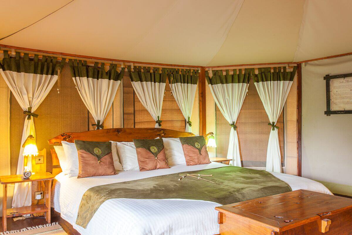 Quarto bem iluminado e espaçoso, com cama grande, vários travesseiros, cabeceira de madeira e cortinas.