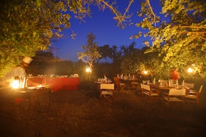 An outdoor dining area with tables and chairs, surrounded by trees and illuminated by string lights.