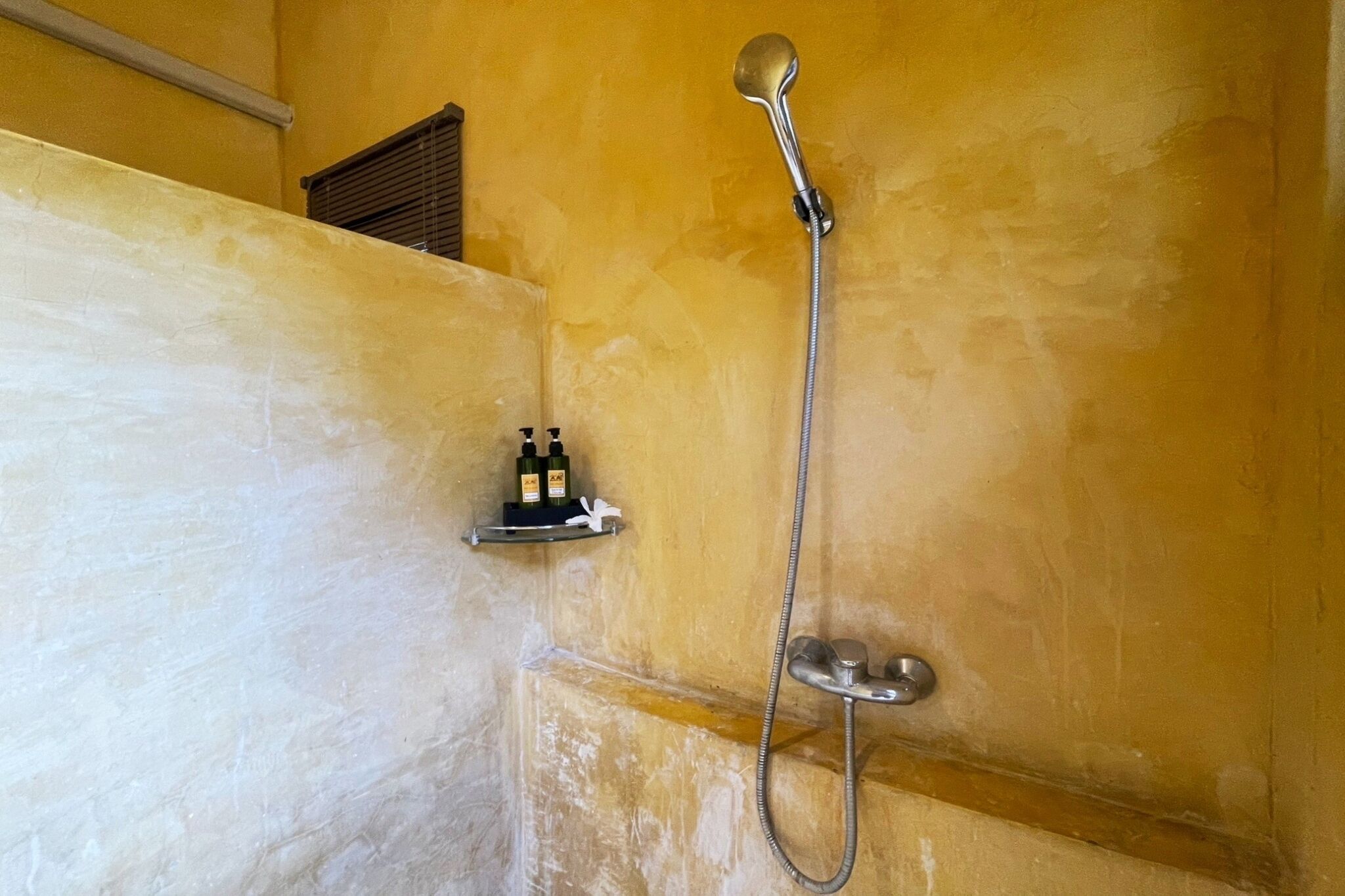 A shower with a handheld showerhead and a wall-mounted faucet. There are two bottles on a shelf beside the shower.