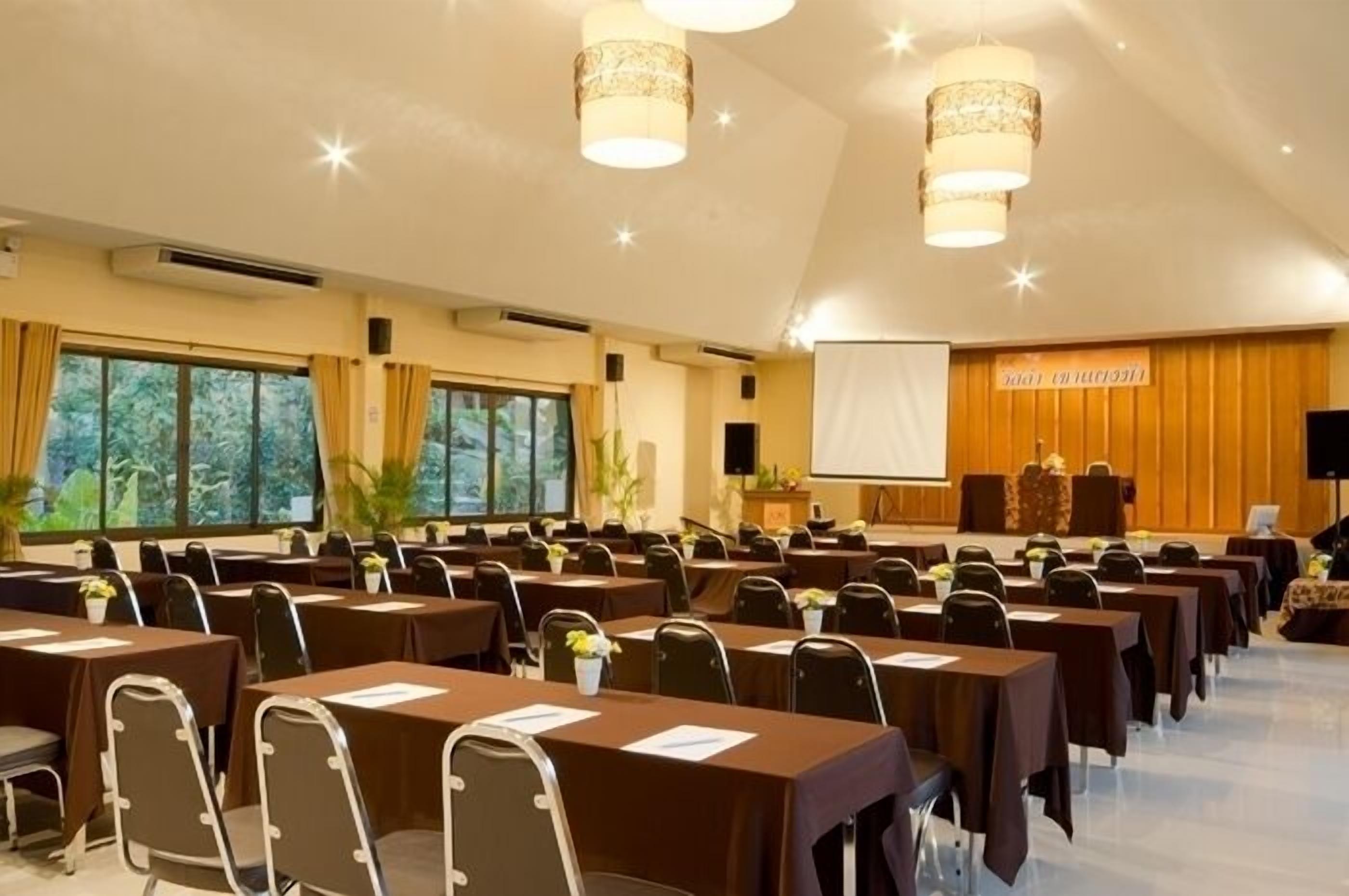 Un salón de conferencias con filas de mesas y sillas, ventanas amplias y un escenario con un atril.