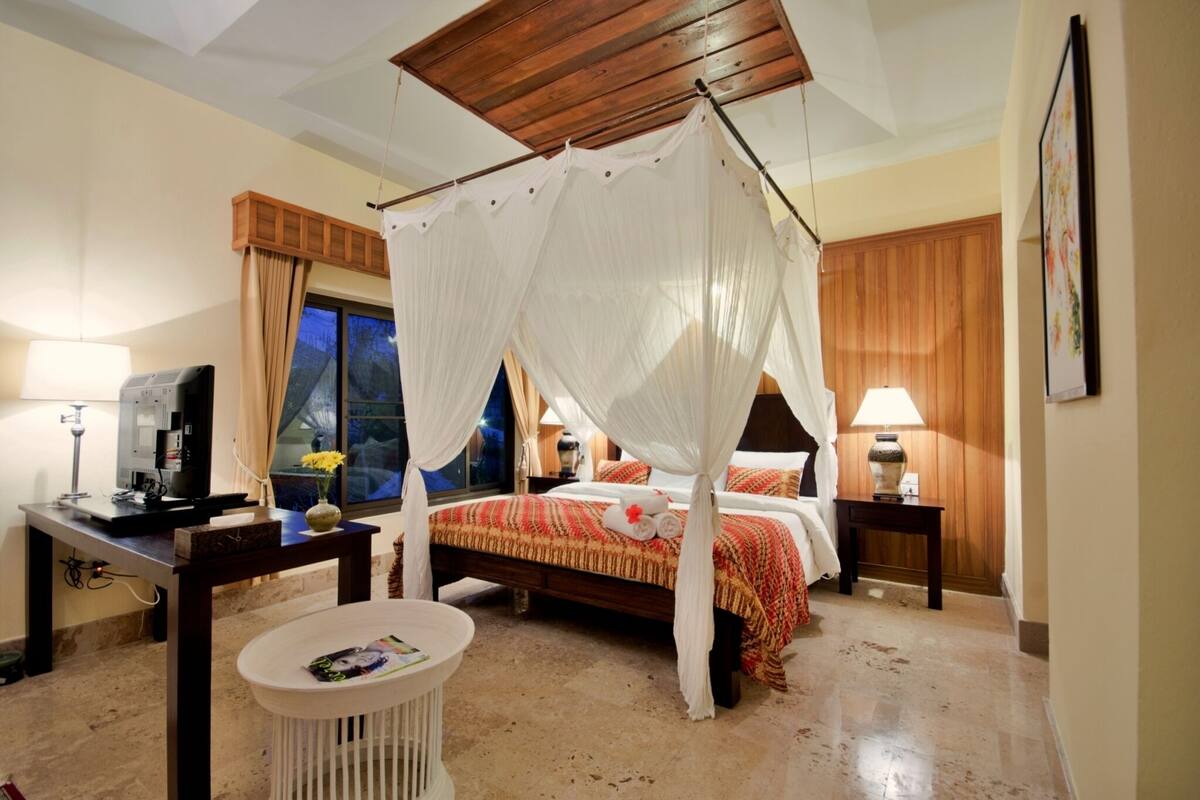 Une chambre d’hôtel avec un lit à baldaquin, une télévision, un bureau avec une lampe et une petite table ronde.