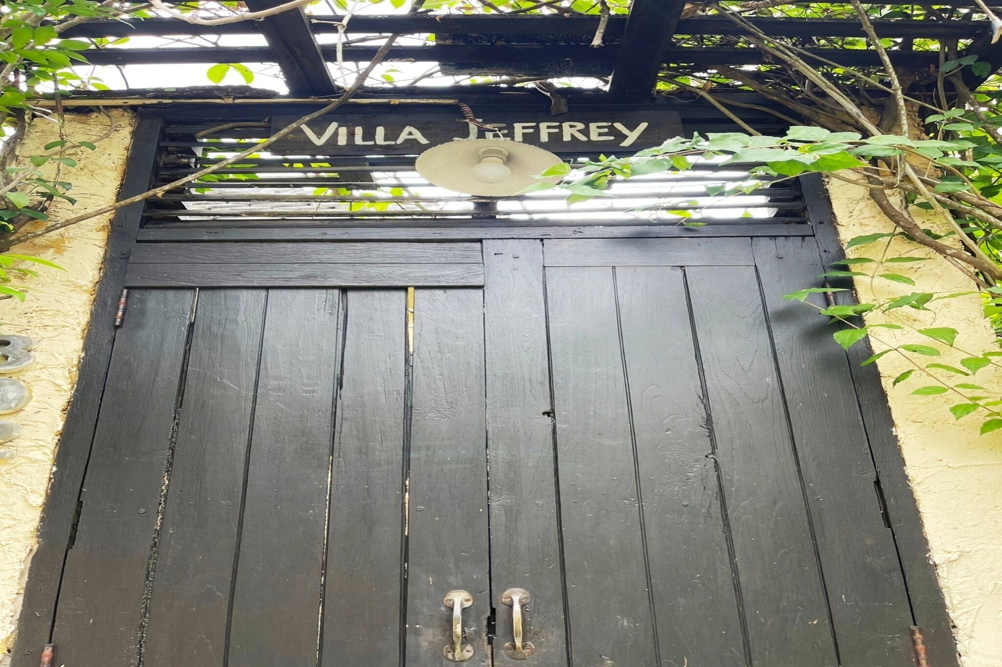 Ein schwarzes Holztor mit der Aufschrift 'Villa Jeffrey' darüber, einem Metall-Türgriff und einem Deckenventilator.