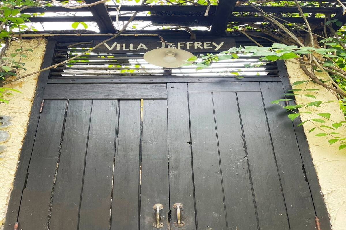 Un portail en bois noir surmonté de l’inscription « Villa Jeffrey », une poignée de porte en métal et un ventilateur de plafond.