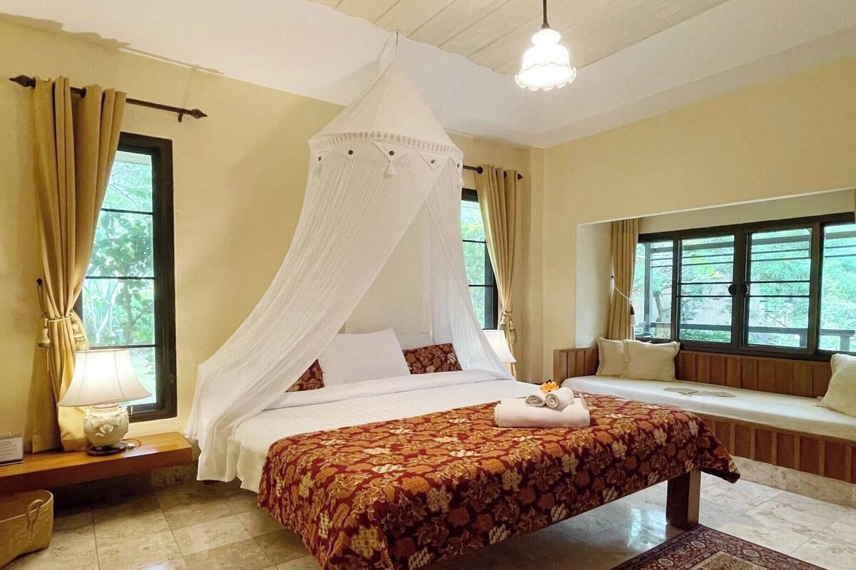 Une chambre à coucher avec un lit à baldaquin, un banc agrémenté d’un coussin et une vue sur la verdure par les fenêtres.