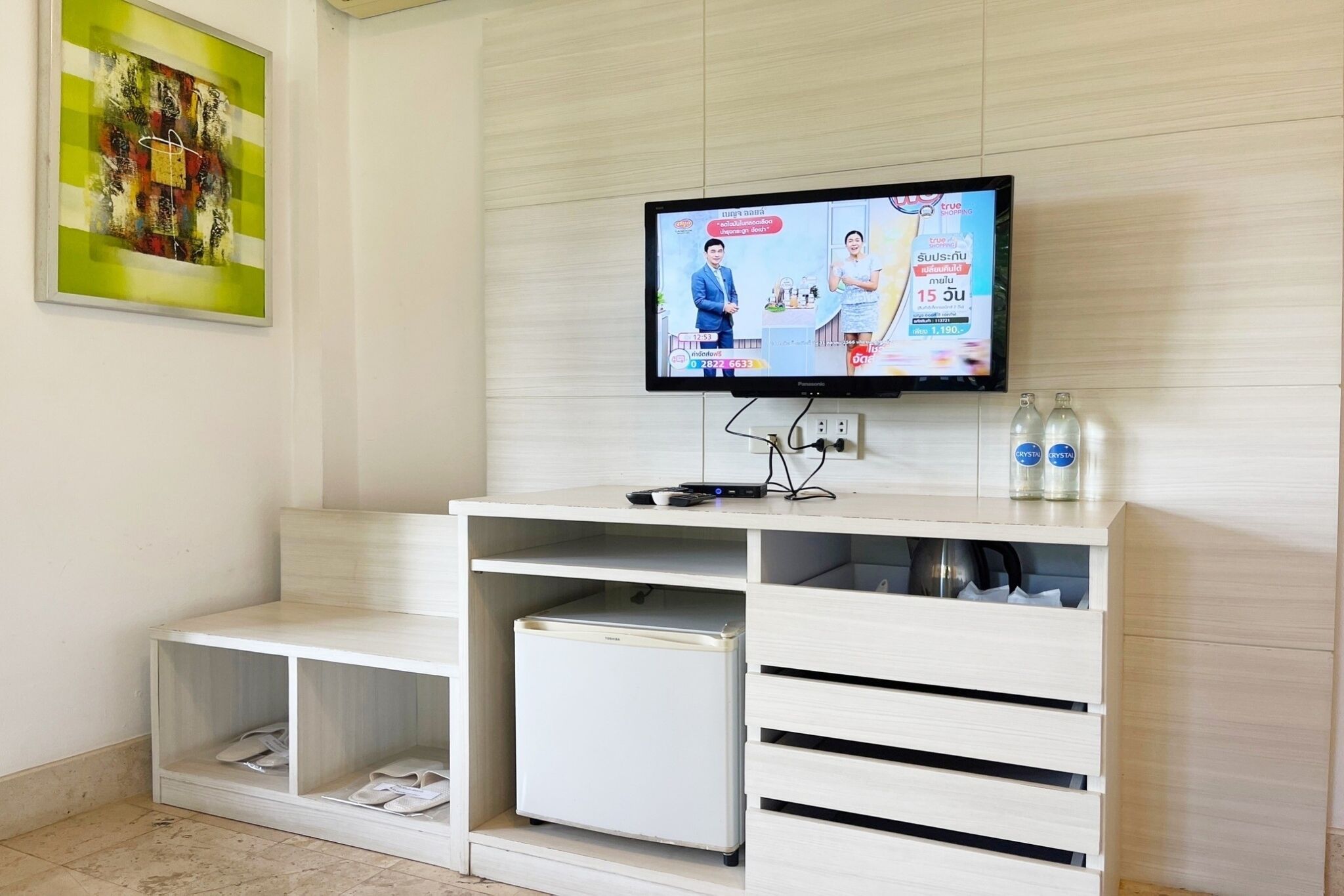A hotel room with a television mounted on the wall, a white desk with drawers, a small refrigerator, and a shelf with shoes.