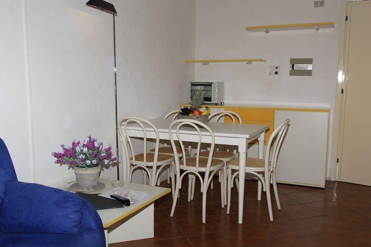 A compact kitchen with a microwave, a small dining table with chairs, a potted plant, and a blue sofa in the foreground.