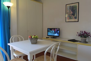 A room with a white table, wicker chairs, a basket of apples, a television, and a framed picture on the wall.