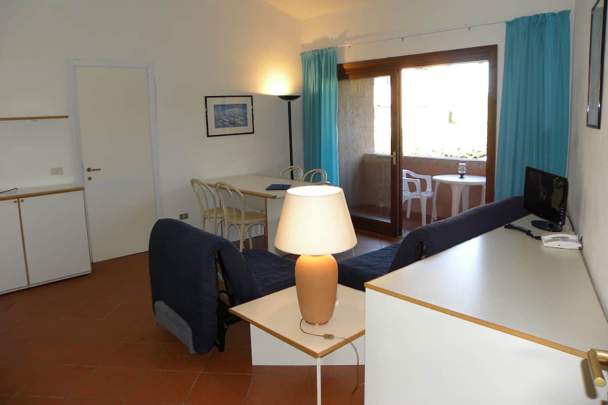 A compact hotel room with a kitchenette, a dining area with a table and chairs, a sofa, and a desk with a lamp.