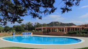 Una piscina amplia y cristalina con un área de terraza techada, rodeada de césped cuidado y árboles.