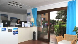 Zona de recepción con mostrador, computadora y carteles informativos. Hay una silla de mimbre y una planta en maceta cerca de la entrada.