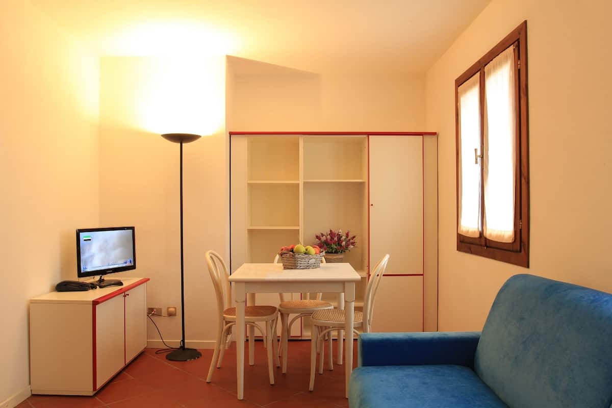 A small room with a white table, four chairs, a blue sofa, a TV on a cabinet, and a floor lamp.