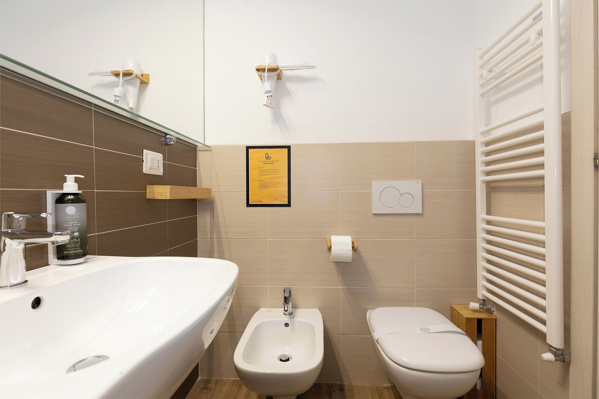 Ein modernes Badezimmer mit einem großen, weißen Waschbecken, einem Bidet und einer Toilette. An der Wand hängt ein informativer Plakatrahmen, ein Handtuchhalter und eine Seifenspender.