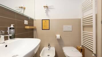 Ein modernes Badezimmer mit einem großen, weißen Waschbecken, einem Bidet und einer Toilette. An der Wand hängt ein informativer Plakatrahmen, ein Handtuchhalter und eine Seifenspender.