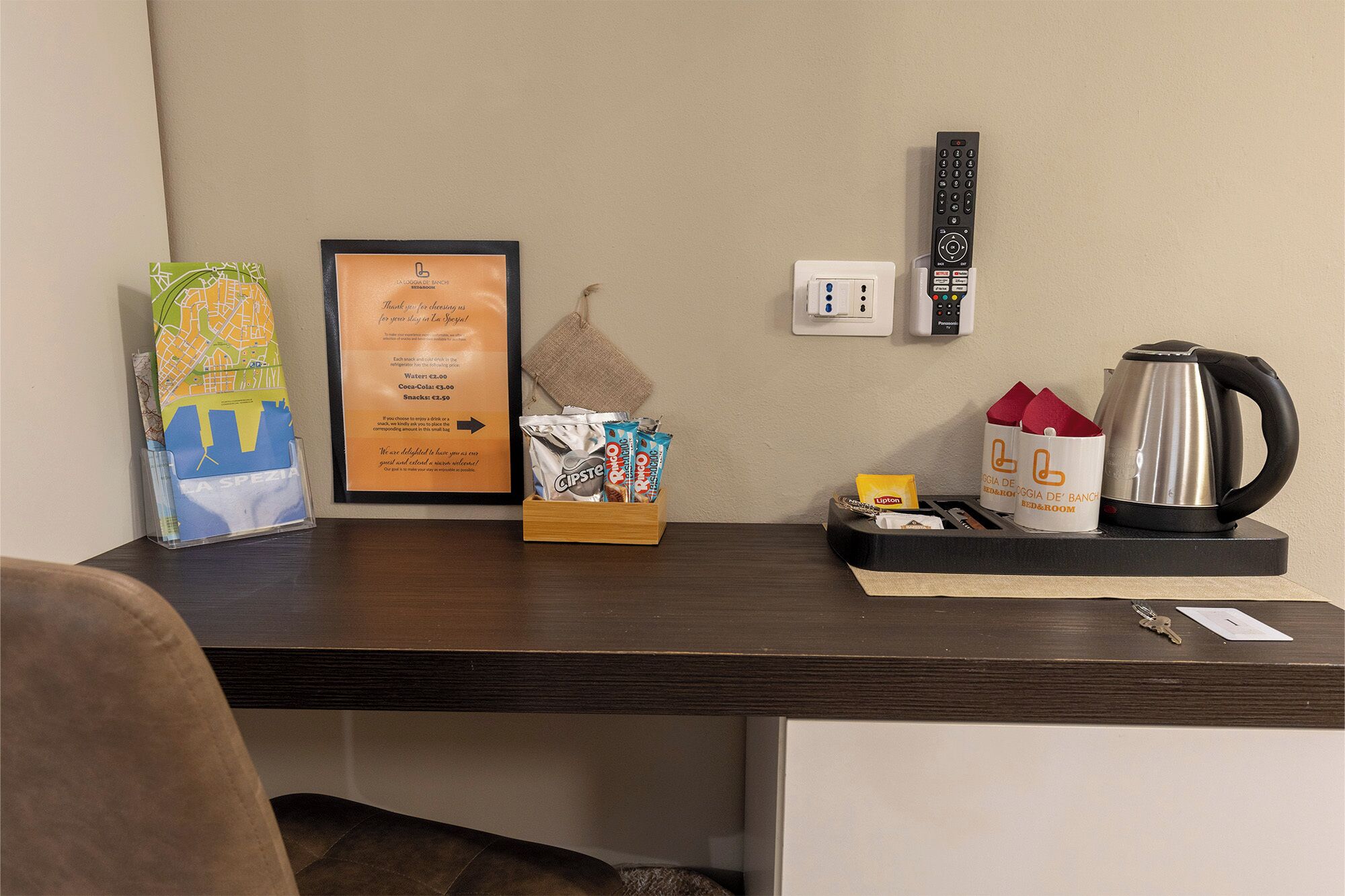 Un bureau avec un menu encadré, une bouilloire, une télécommande et un petit plateau contenant des serviettes en papier et une tasse.