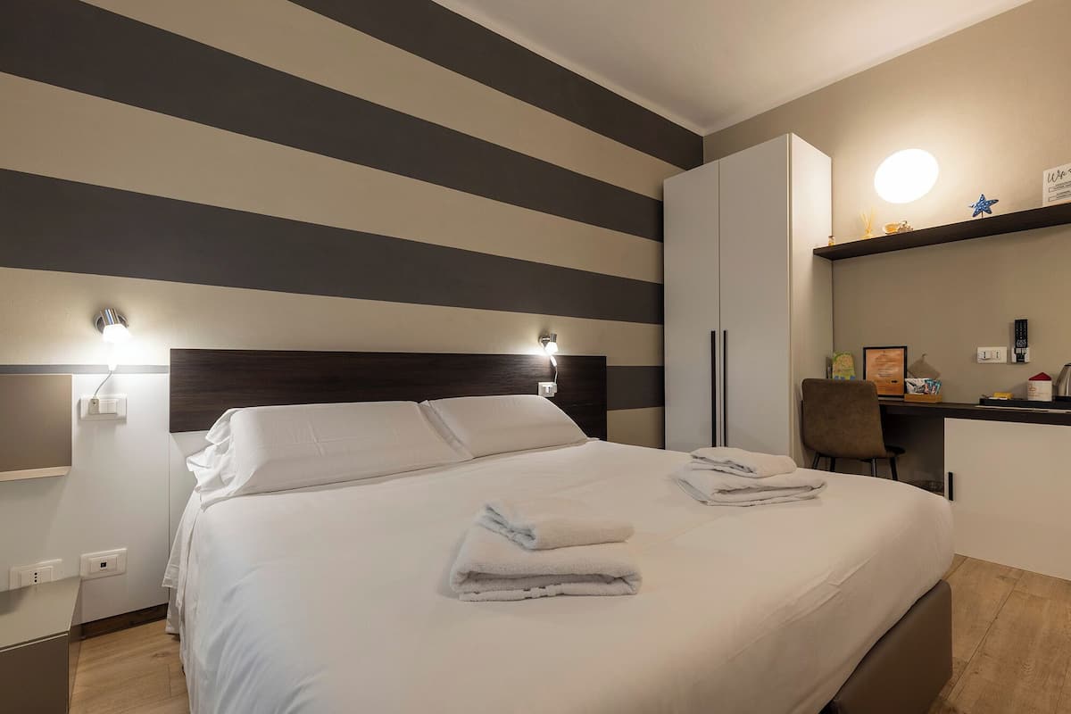 A hotel room with a bed, bedside lamps, a desk with a chair, and a shelf with items.