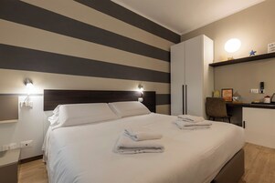 Una habitación de hotel con cama, lámparas de noche, un escritorio con silla y estante con objetos.