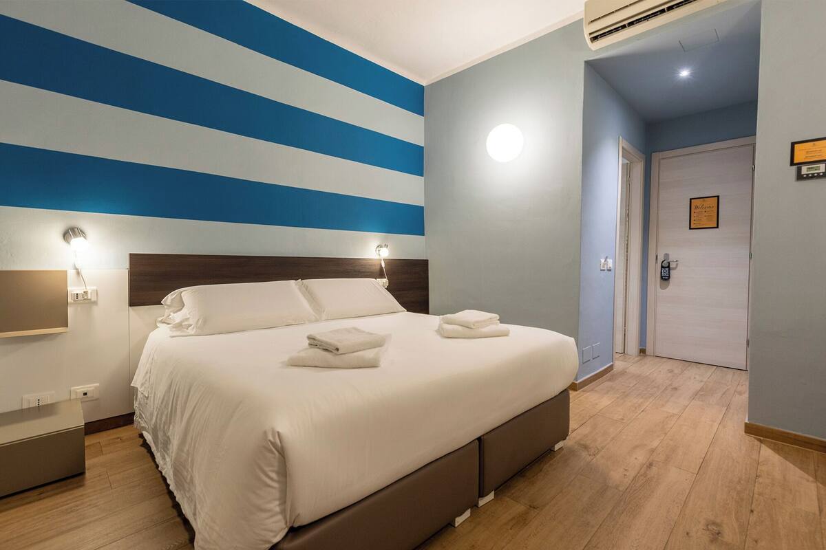 A hotel room with a bed, bedside tables, a wall with blue stripes, a door, and a light fixture.