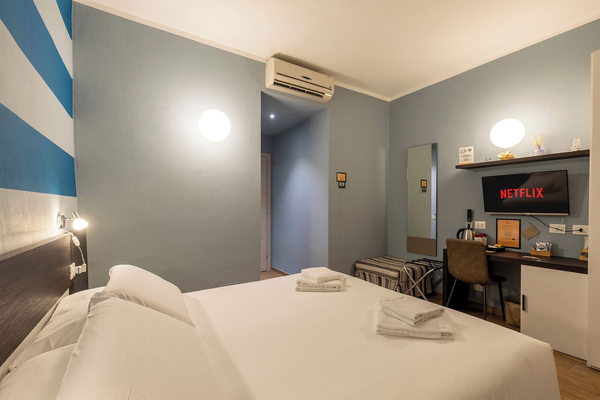 Una habitación de hotel con una cama, un escritorio con una computadora, un televisor mostrando 'NETFLIX' y un equipo de aire acondicionado.