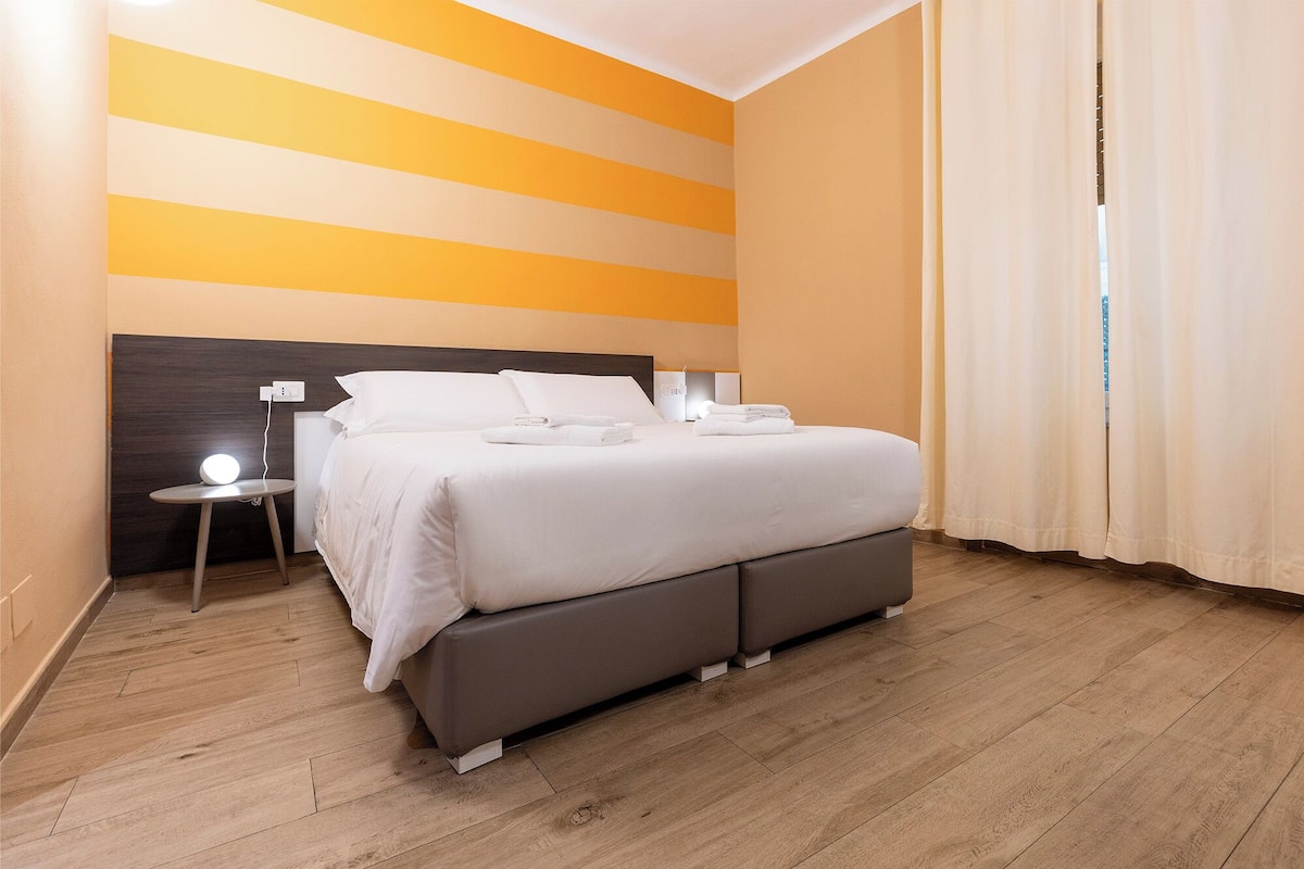 A modern hotel room with a large bed, a bedside table with a lamp, and a view of the outdoors through the window.