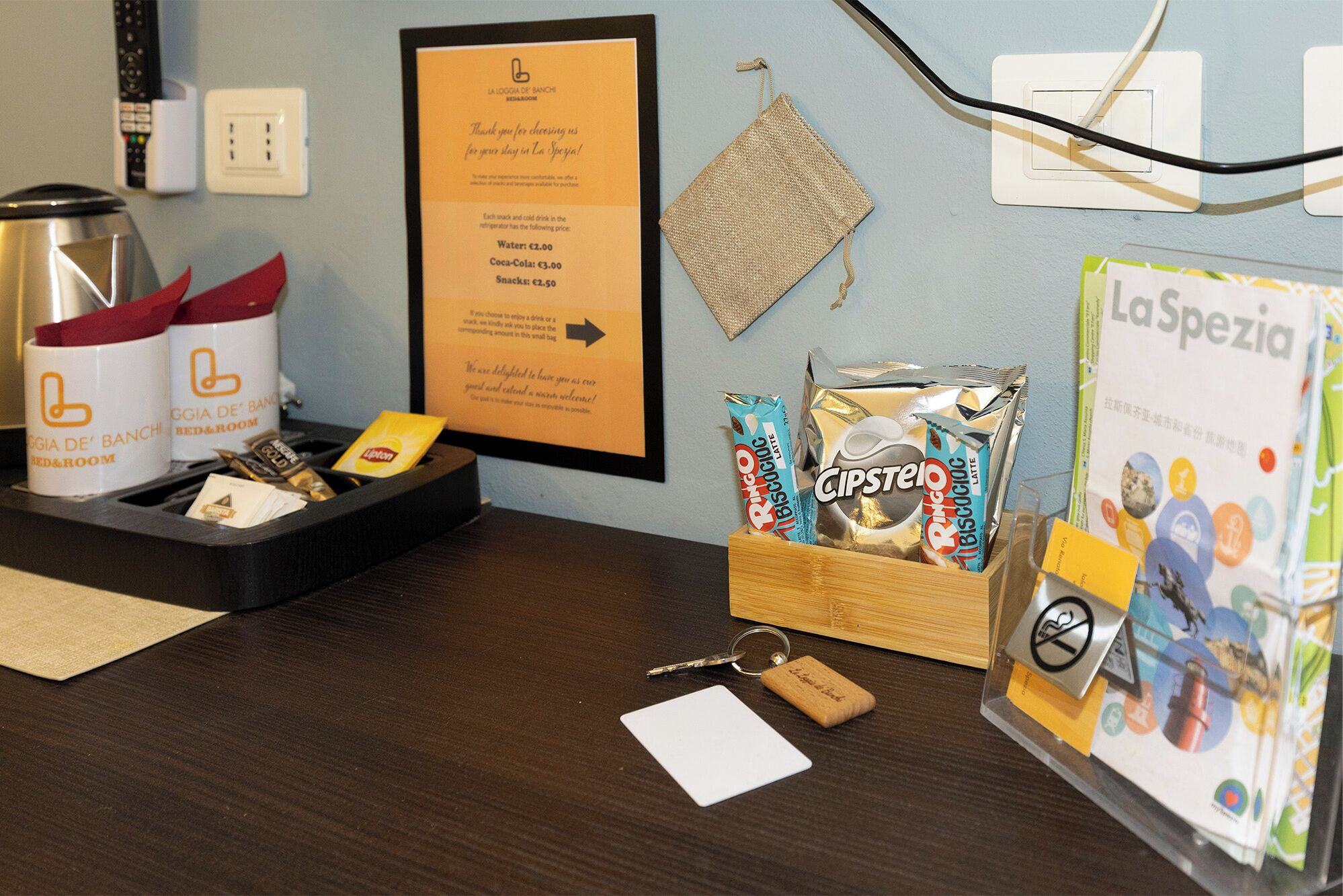 Un mostrador de recepción de hotel con un mensaje de bienvenida enmarcado, un mini bar con aperitivos y bebidas, un dispensador de tarjetas de llave y una revista de La Spezia.