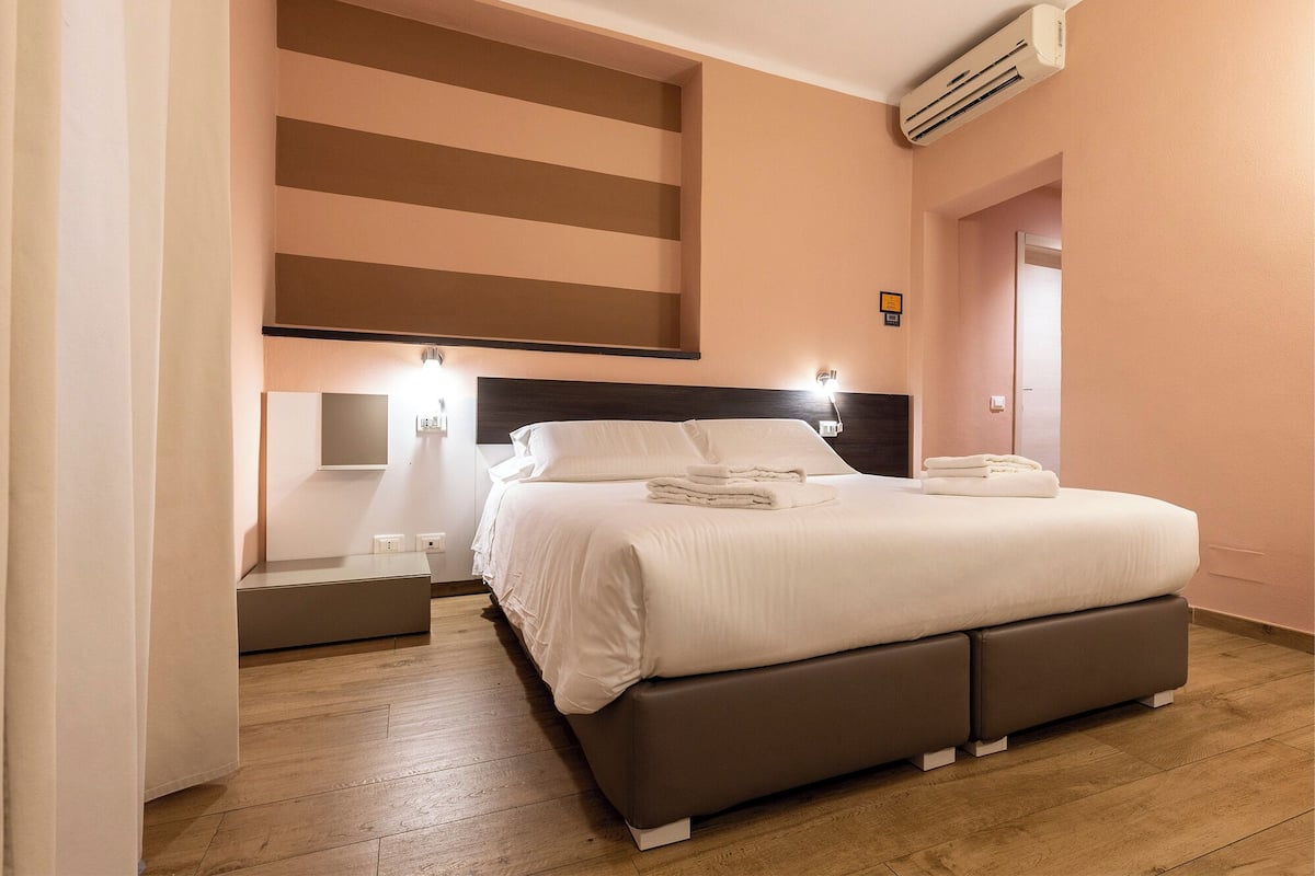 A modern hotel room with a large bed, bedside tables, a wall-mounted air conditioner, and a wooden floor.