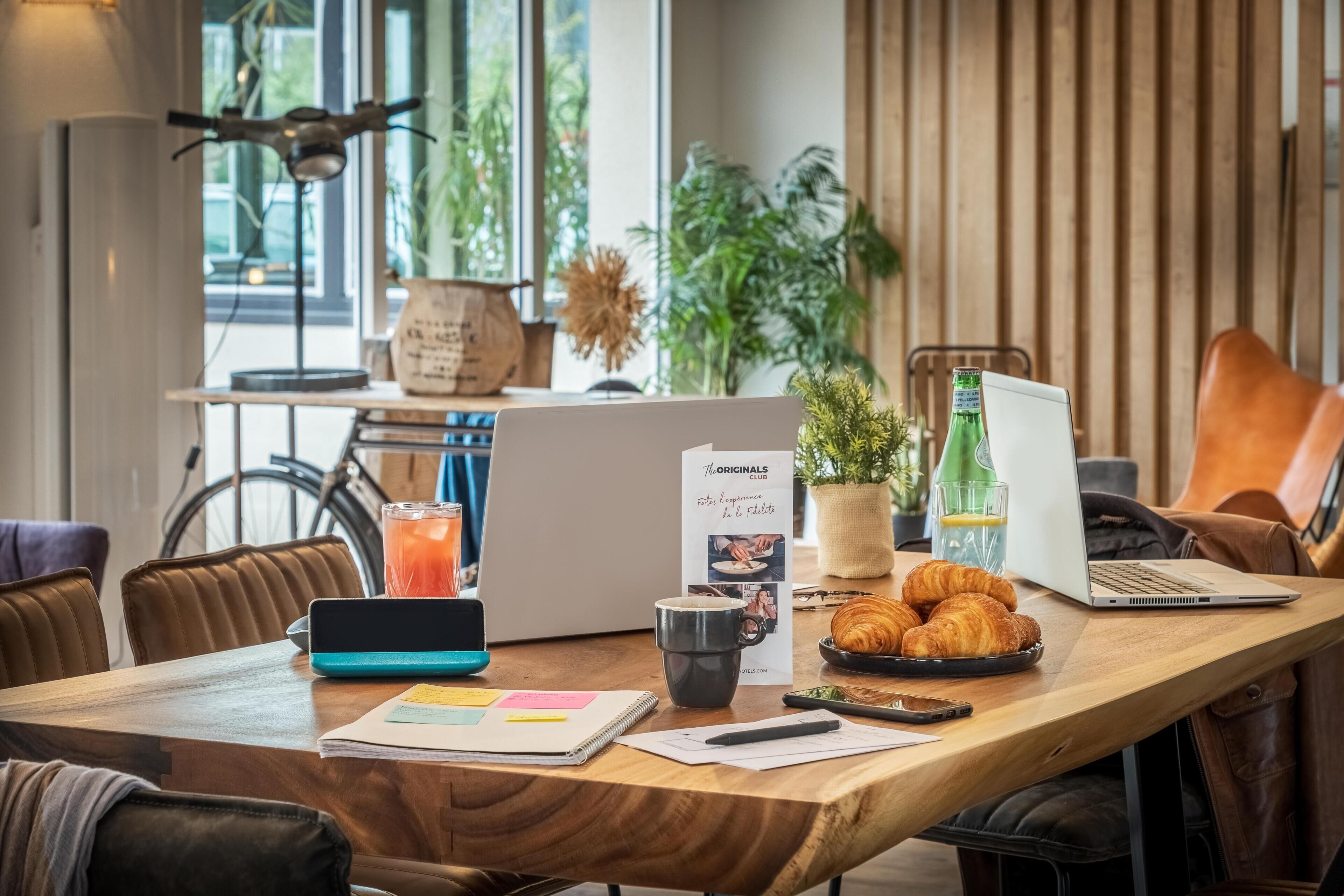 Une table en bois sur laquelle se trouvent un ordinateur portable, un carnet, un téléphone intelligent, une tasse, une assiette de croissants et un menu.