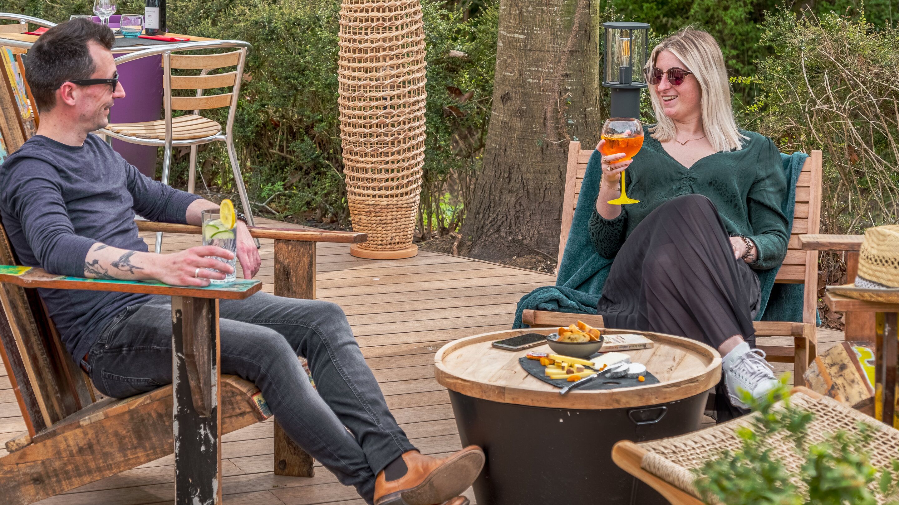 A couple seated at an outdoor wooden deck with a round table, surrounded by wicker furniture and a small garden.