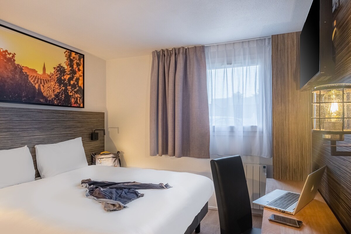 A hotel room with a large bed, a desk with a laptop, a desk lamp, and a painting on the wall.