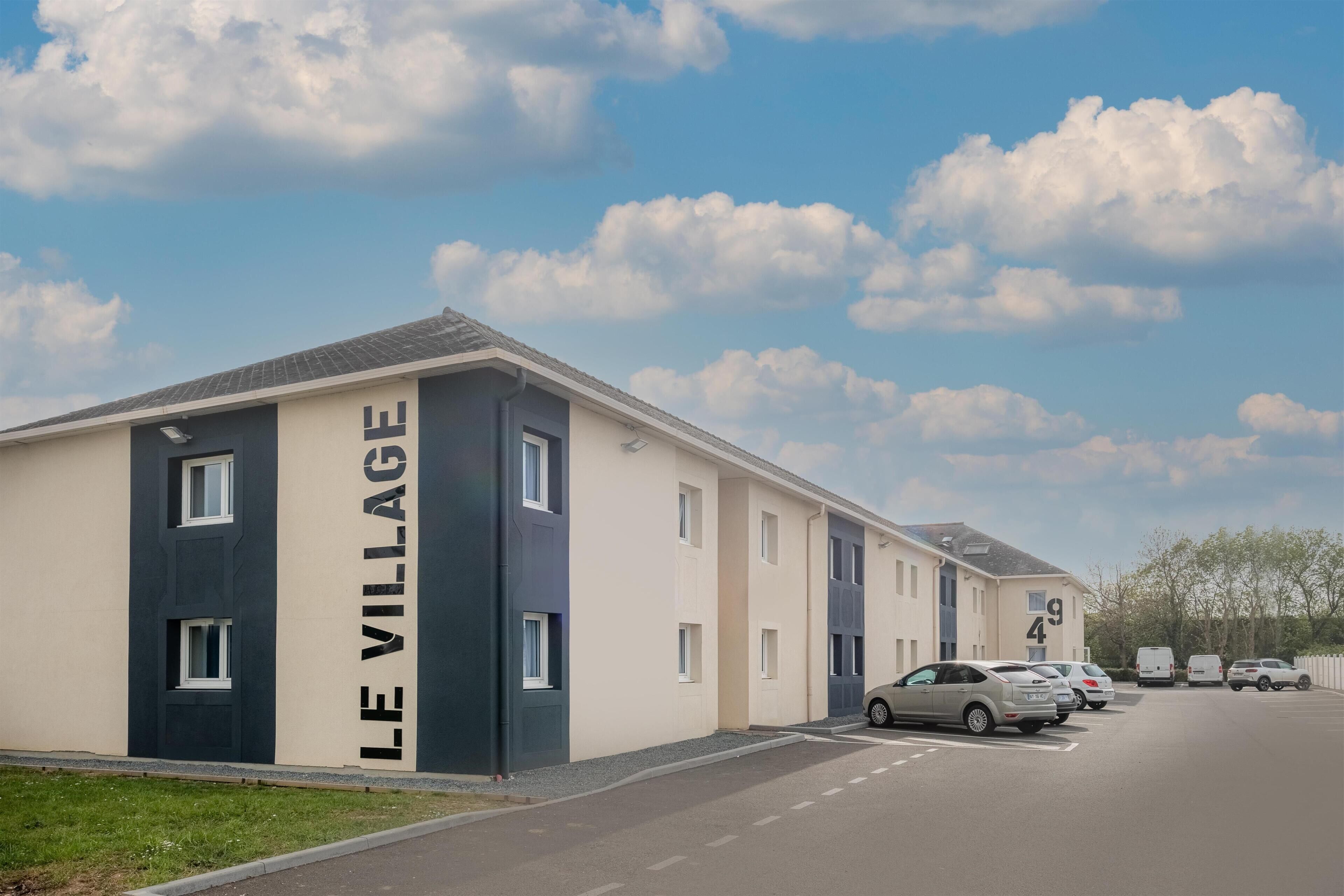 A modern building with 'LE VILLAGE' signage, a parking lot with several cars, and a clear sky.