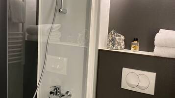 Un baño con cabina de ducha de vidrio, toallero, estante con artículos de aseo y un interruptor de luz en la pared.