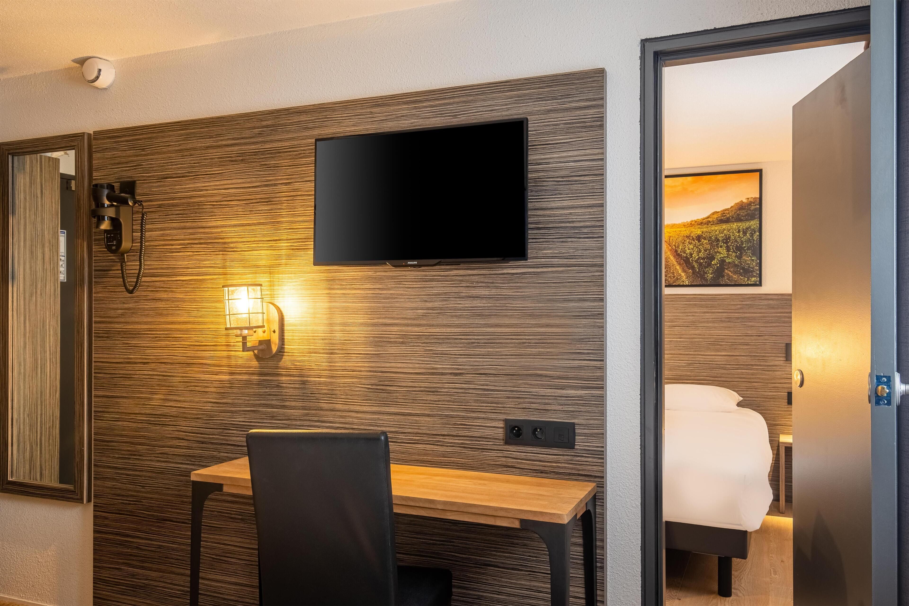 Une chambre d’hôtel équipée d’un téléviseur à écran plat fixé au mur, d’un bureau avec une chaise, d’un lit et d’un tableau accroché au mur.