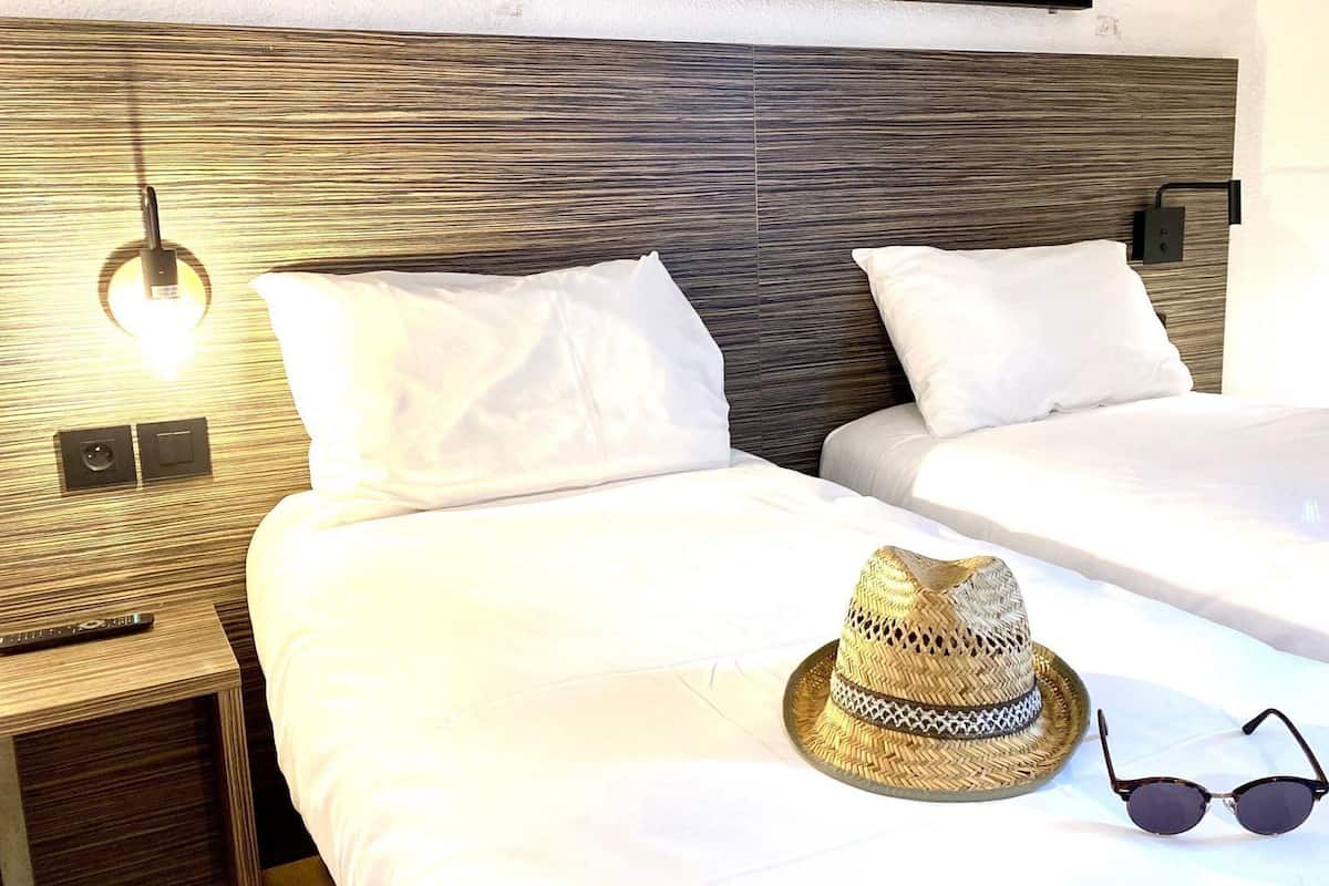 Une chambre d’hôtel avec deux lits, une table de chevet avec une lampe, et un chapeau tressé et des lunettes de soleil posés sur le lit.