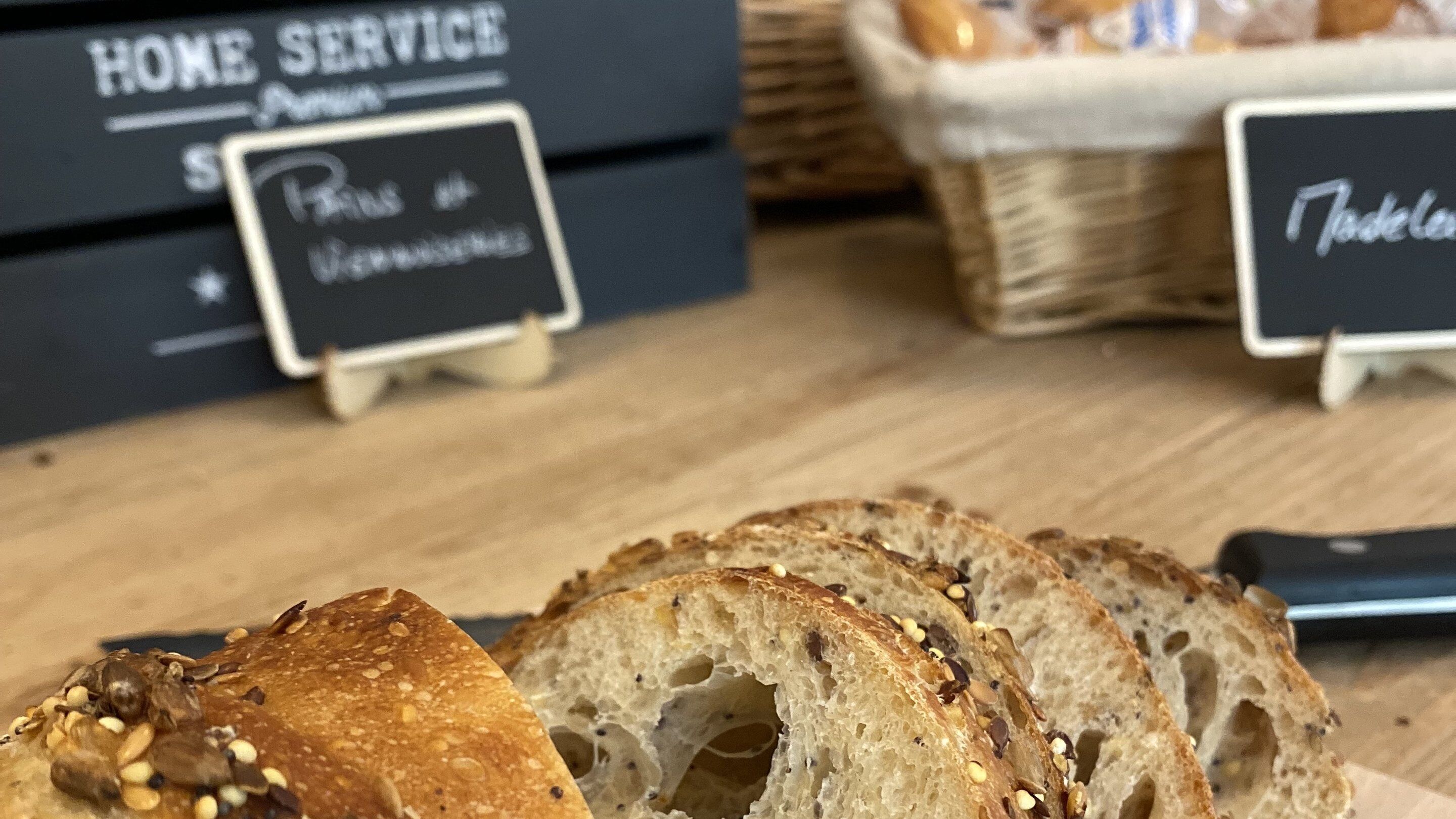 Brot mit Samen auf einem Holztisch, im Hintergrund ein Schild mit der Aufschrift 'HOME SERVICE'.