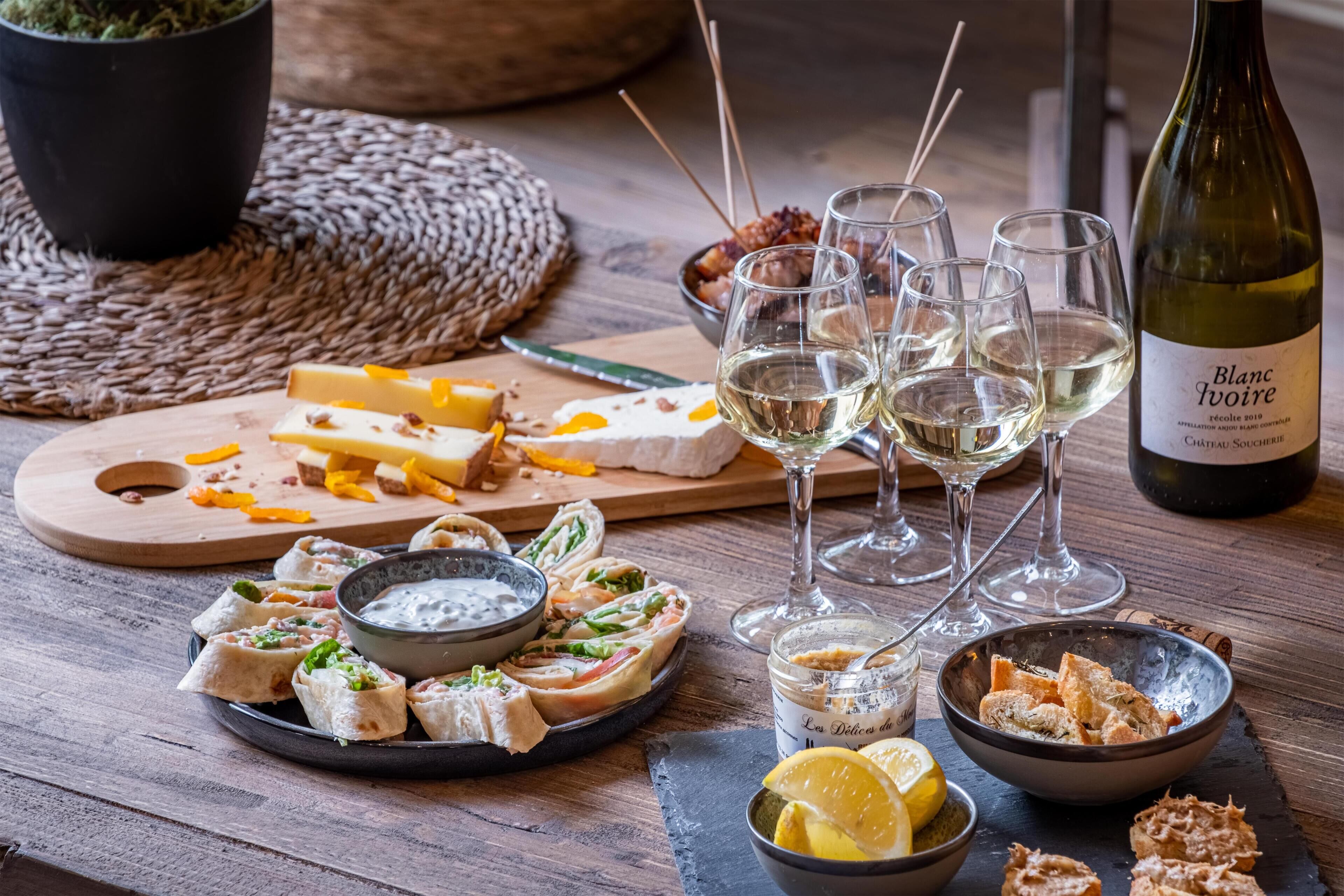 Una mesa de madera con diversos alimentos, incluyendo queso, pan y salsas, junto con varias copas de vino blanco y una botella de Blanc Ivoire.