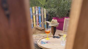 Eine Holzterrasse mit einem runden Tisch, einem Korbstuhl, einem Strohhut, einem Getränk mit Zitronenscheibe und einem Buch.
