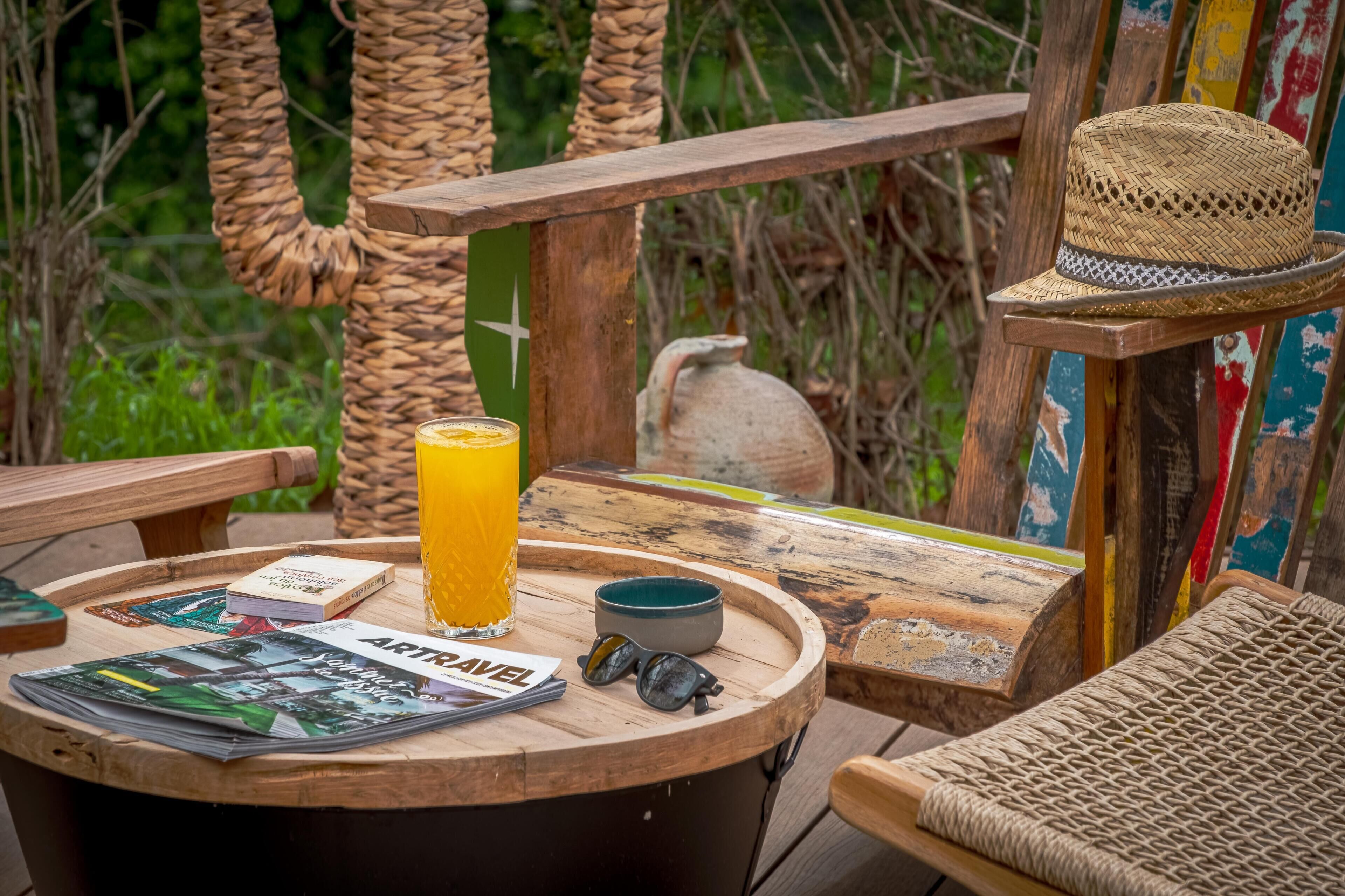 Una terraza de madera con una mesa redonda, un vaso de jugo de naranja, un cuenco y un sombrero.