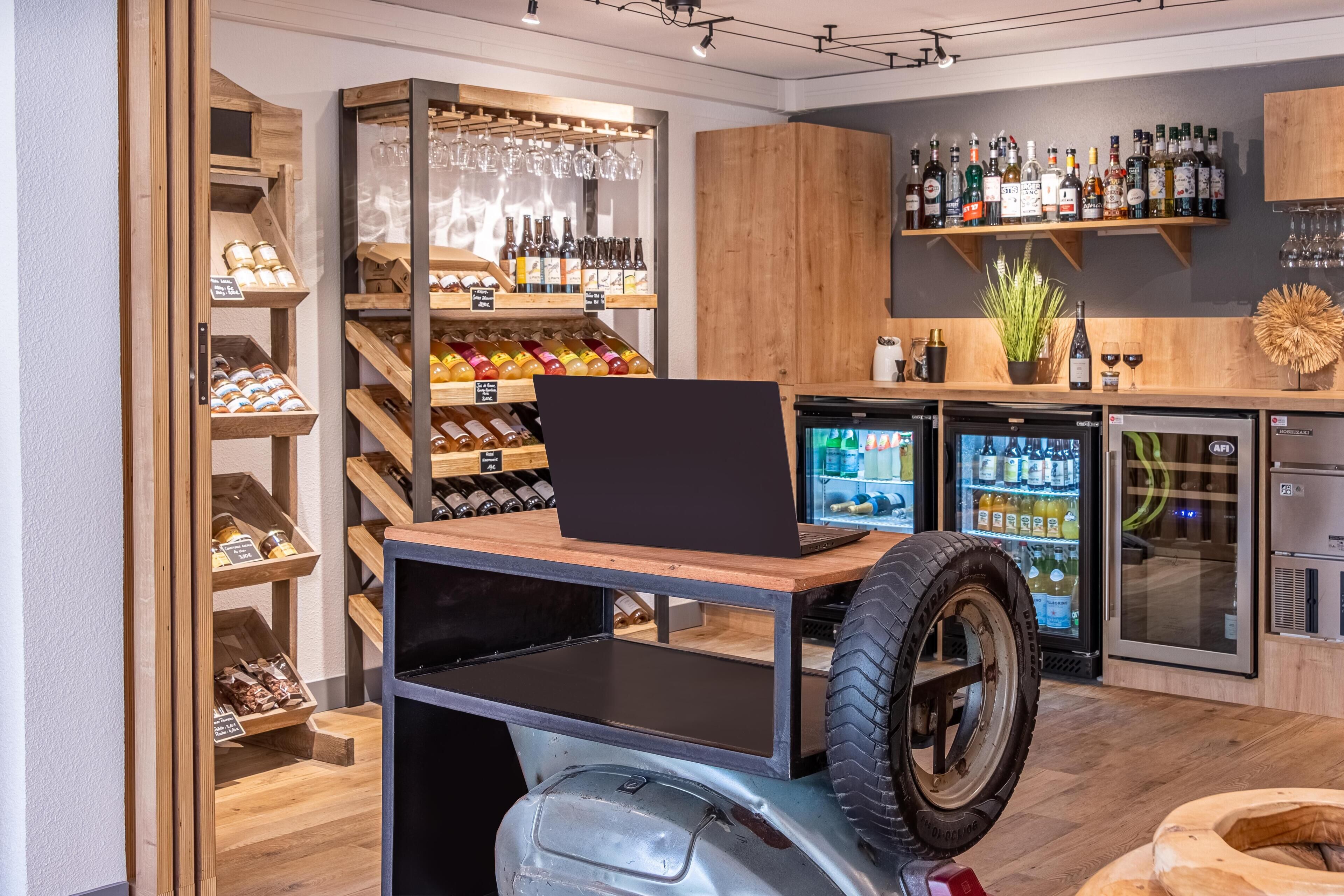 Un espace bar moderne avec un ordinateur portable posé sur un chariot en bois, des étagères remplies de bouteilles d’alcool et un réfrigérateur contenant des boissons.