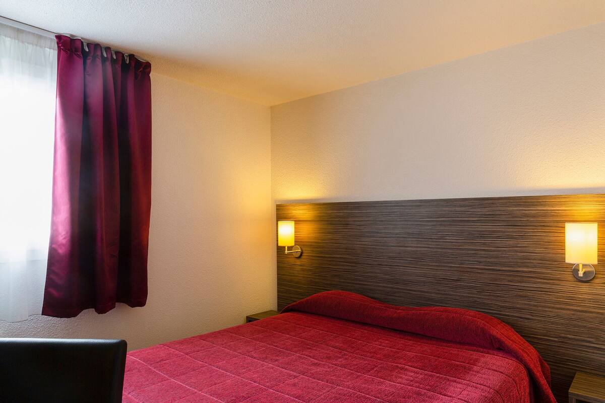 A hotel room with a bed, a nightstand, a lamp, and a window with curtains.