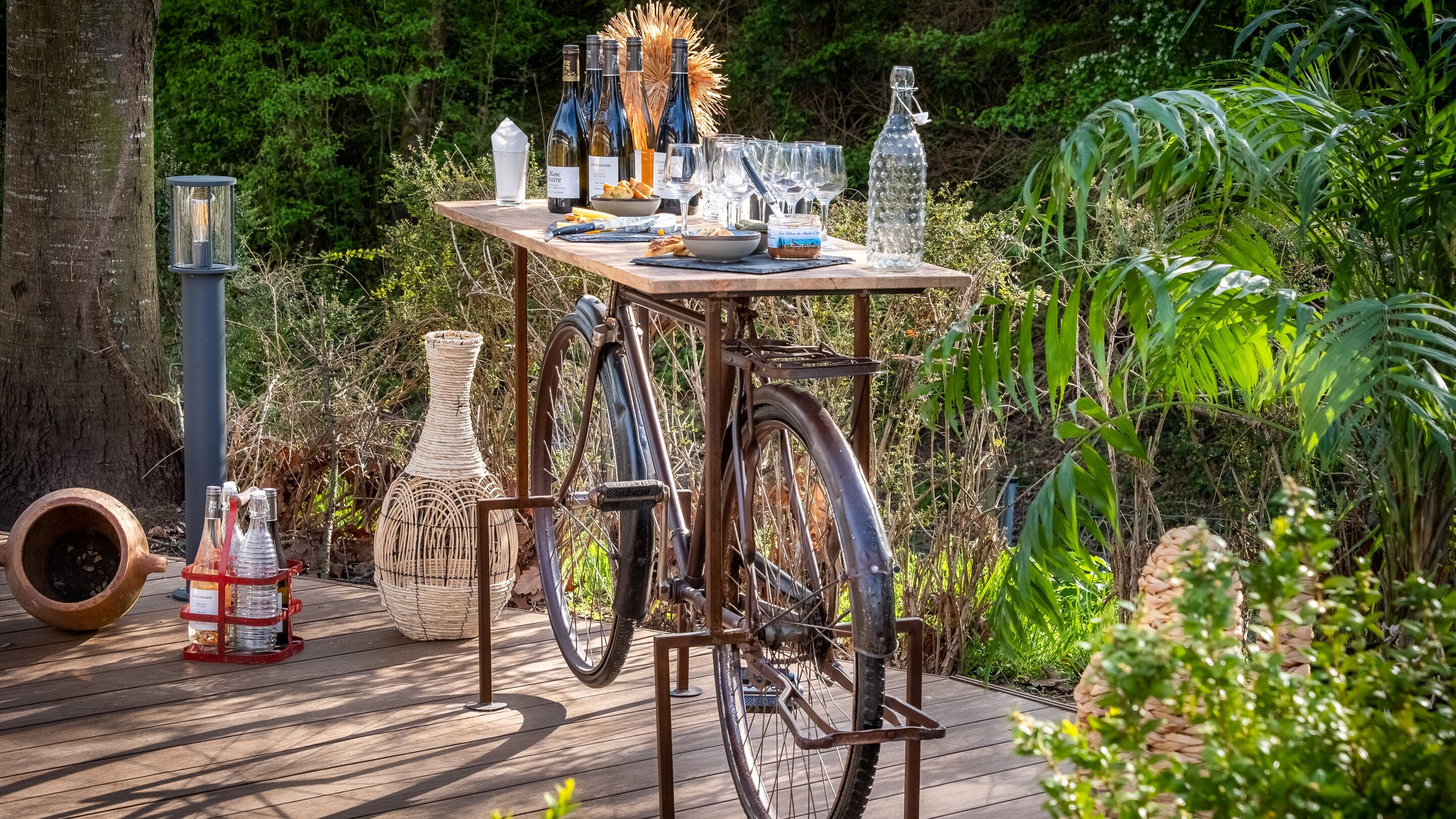 A wooden table set outdoors with wine bottles, glasses, and a wicker basket.