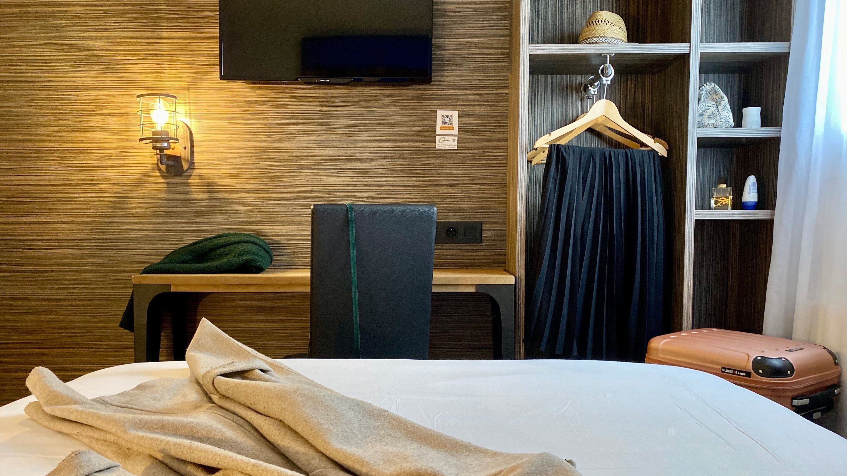 Una habitación de hotel con cama, escritorio, silla, televisor montado en la pared, armario con perchas y maleta.