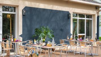 An outdoor dining area with tables and chairs, a large potted plant, and bottles of wine.