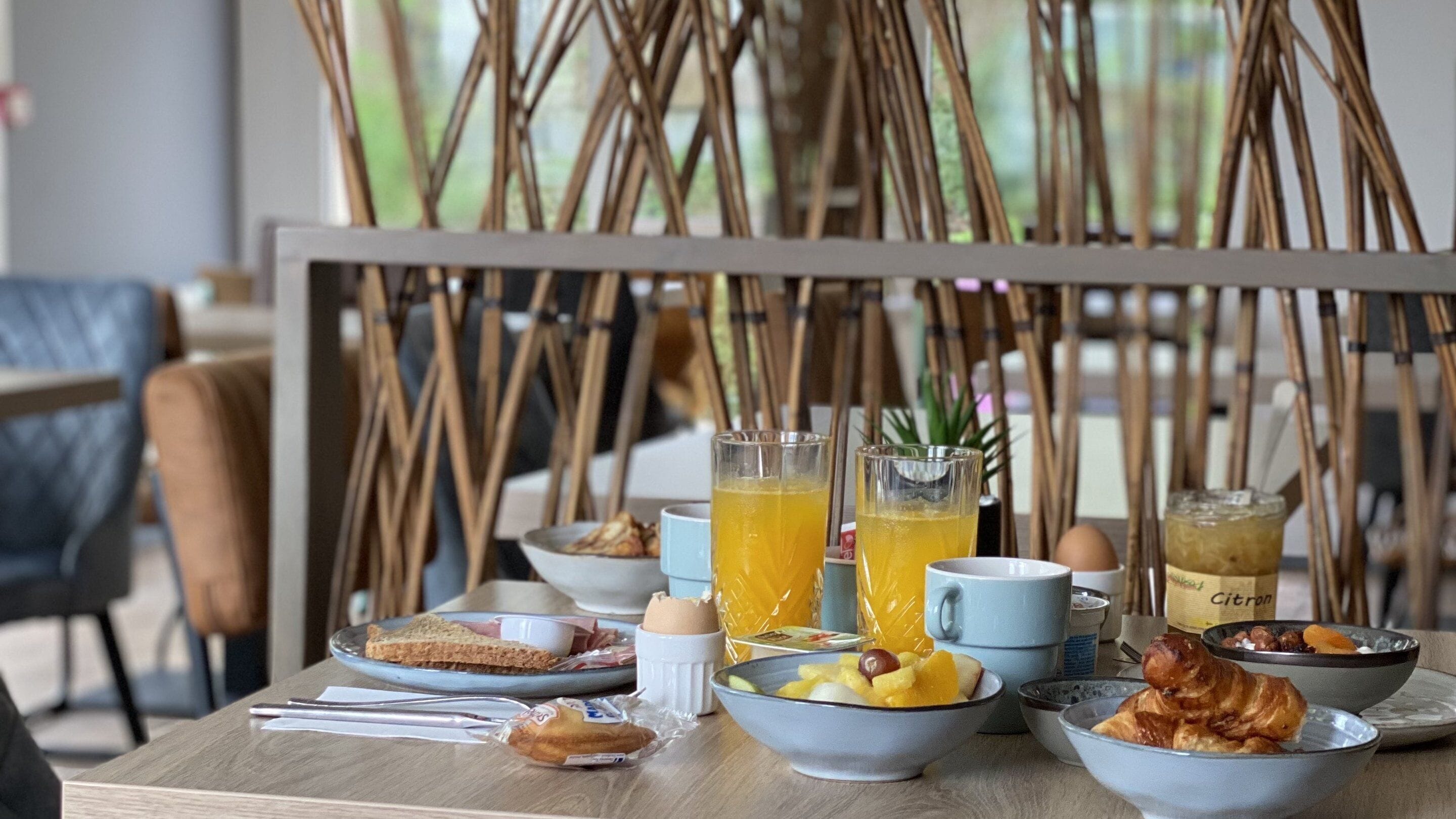 Una mesa de desayuno con varios platos, incluyendo tostadas, fruta y un croissant, junto con dos vasos de zumo de naranja y una botella de jugo.