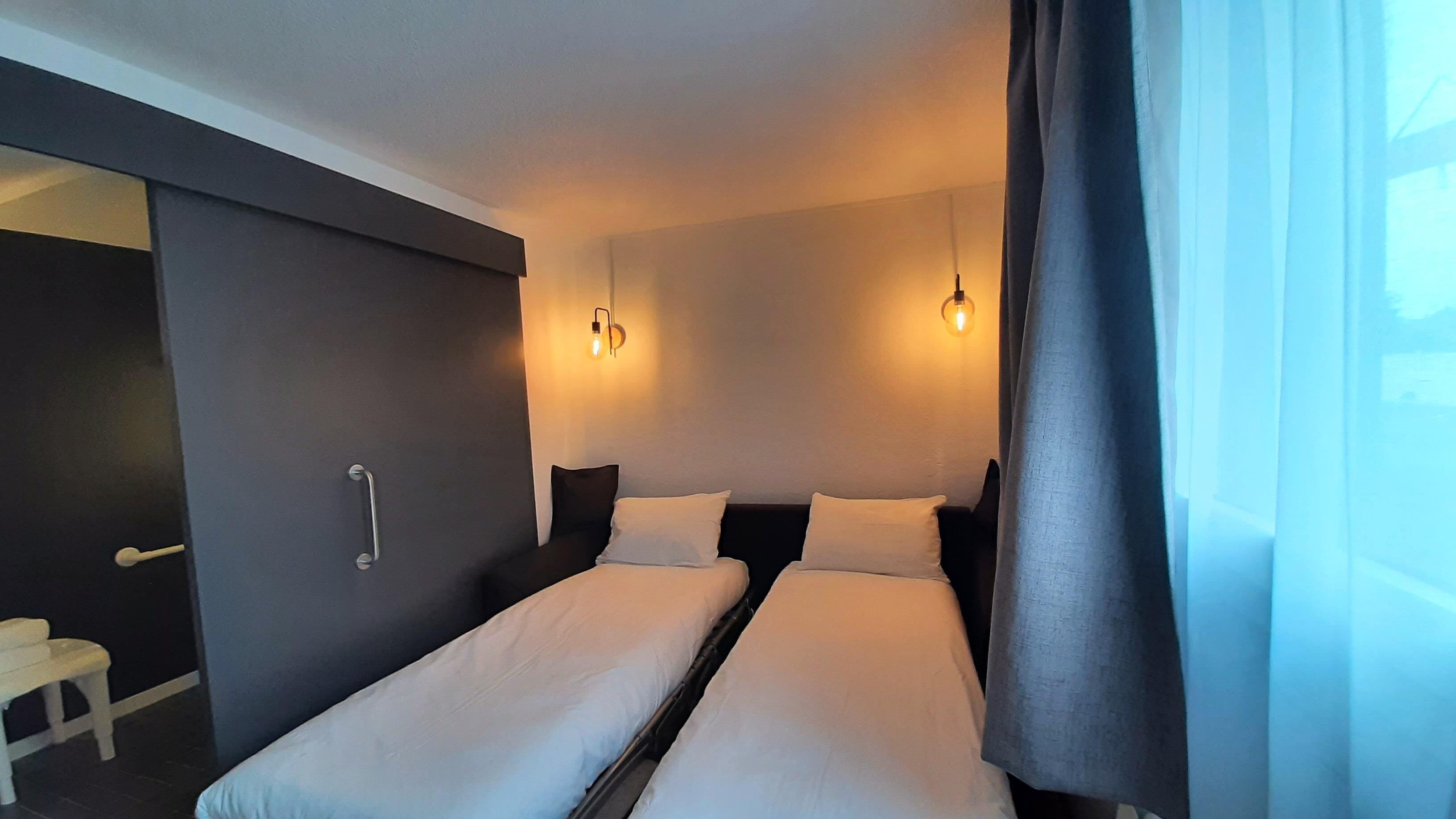 Habitación de hotel con dos camas, una mesita de noche, una silla y una ventana con cortinas.