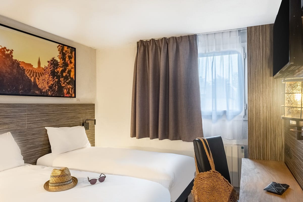 Une chambre d’hôtel avec un lit, un téléviseur fixé au mur, une fenêtre avec des rideaux, une lampe fixée au mur et un tableau accroché au mur.