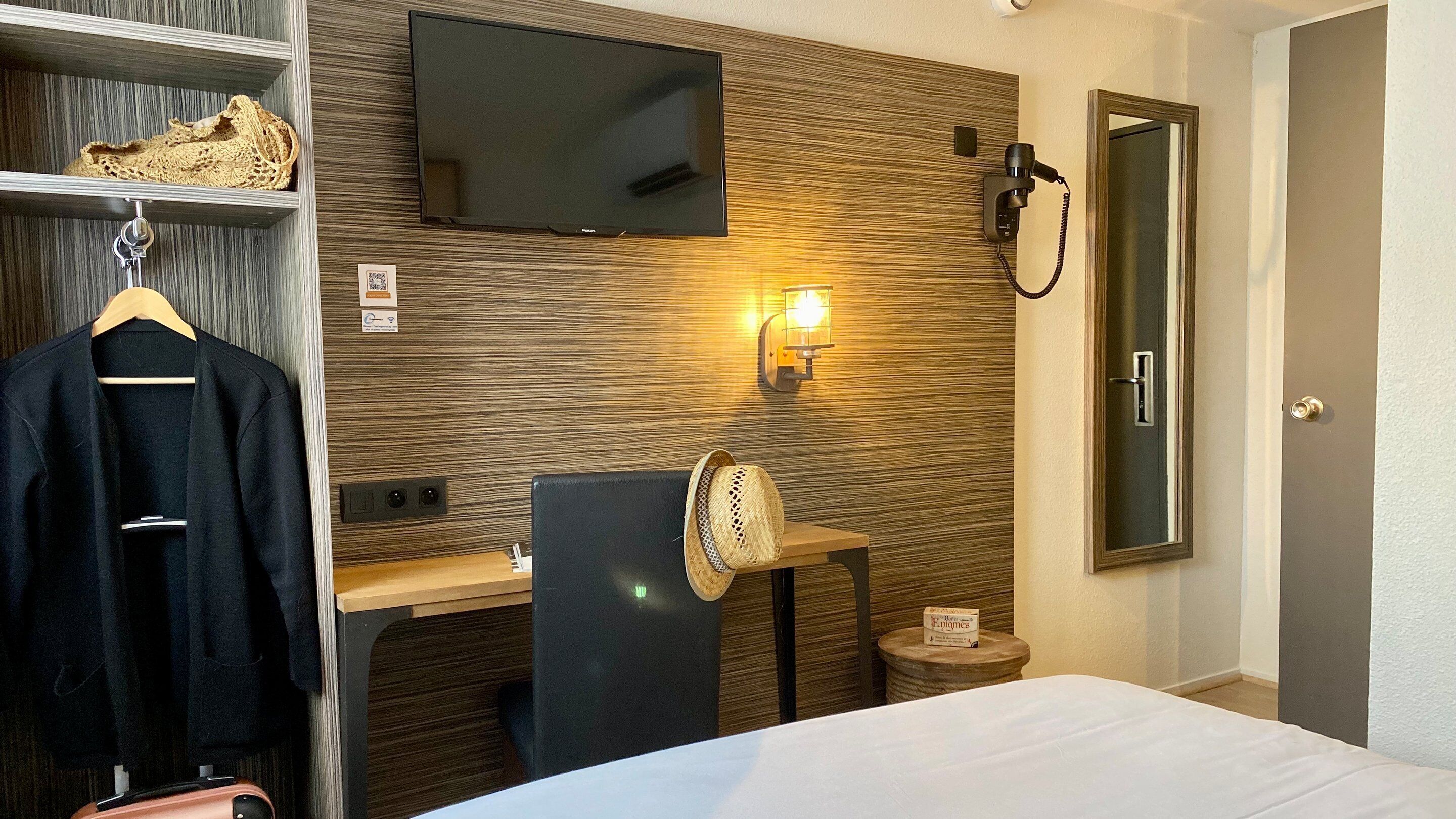 A hotel room with a flat-screen TV mounted on the wall, a desk with a chair, a bedside table with a lamp, a mirror, and a wardrobe with a hanging coat.