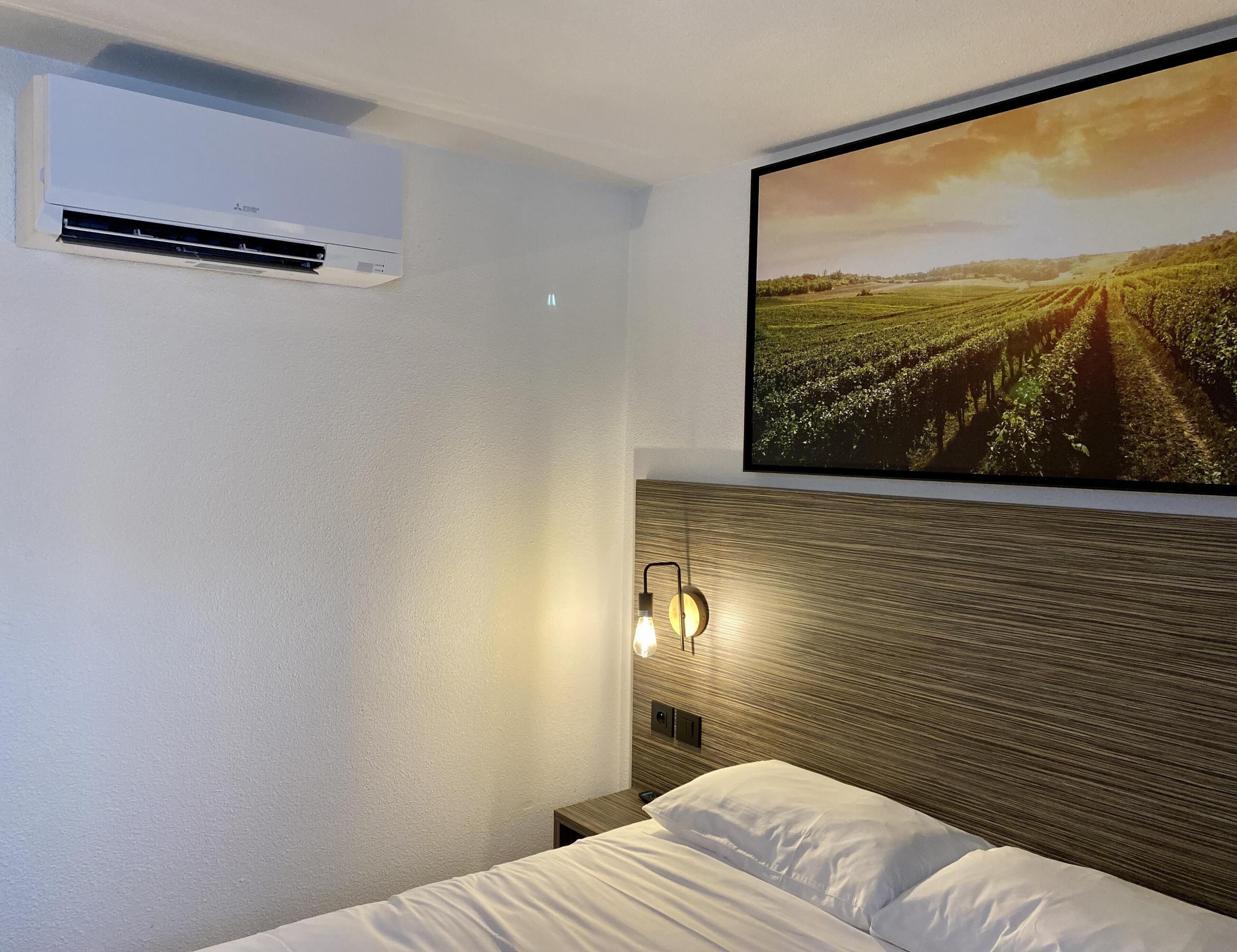 Habitación de hotel con cama, aire acondicionado, lámpara en la pared y un cuadro grande de un viñedo.