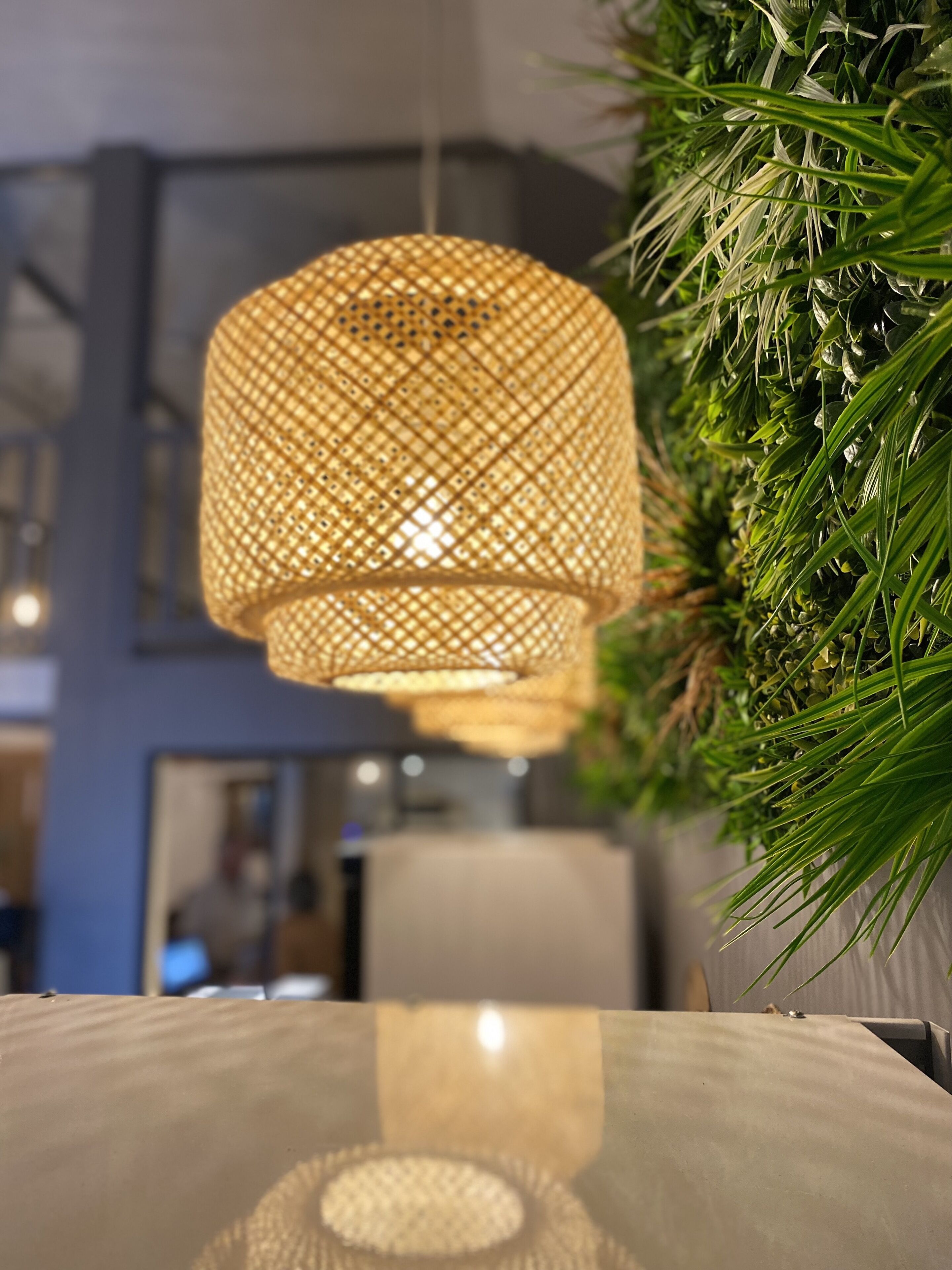 A hanging woven light fixture with a geometric pattern, a glass table reflecting the light, and a green plant with long leaves to the right.