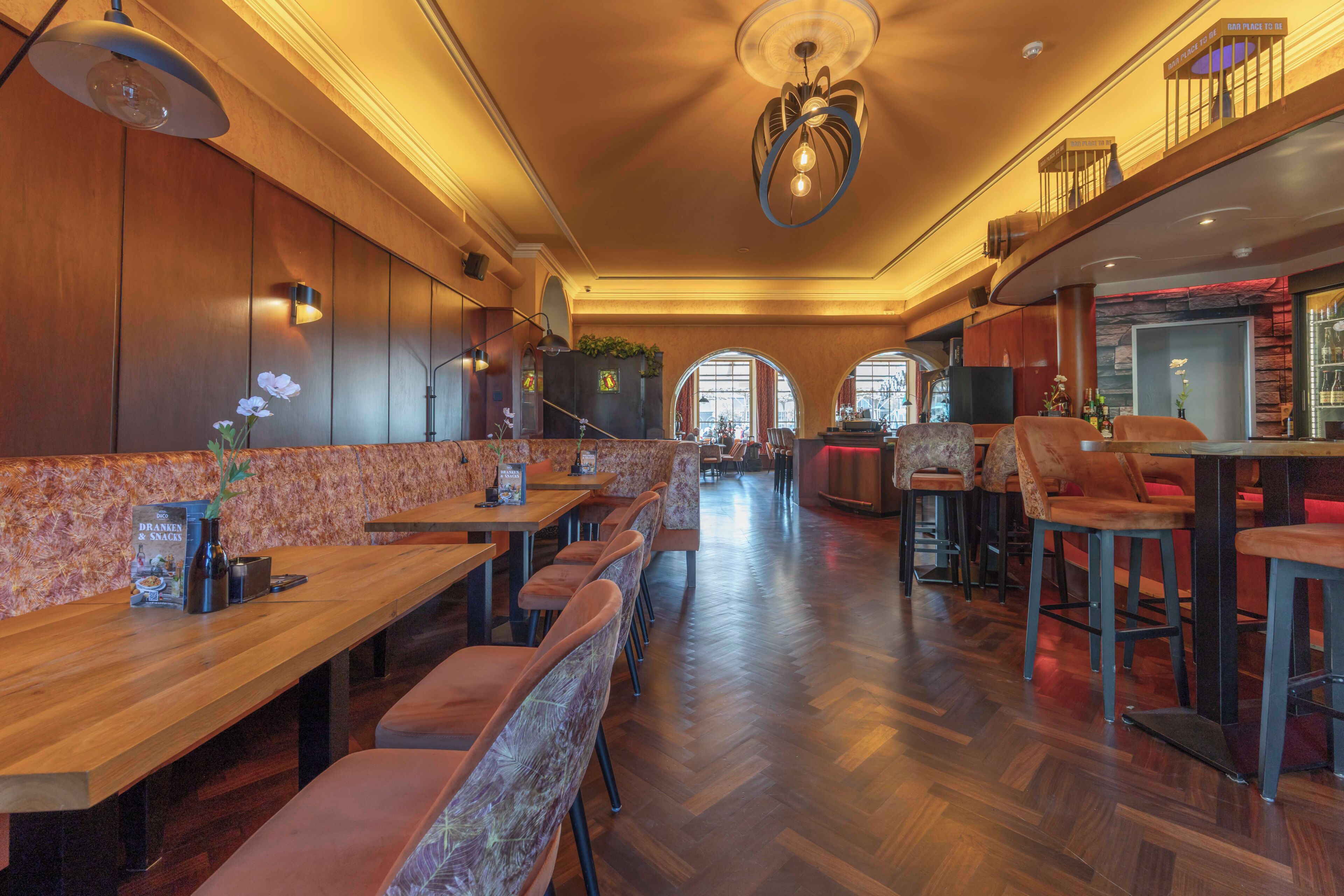 A restaurant with wooden tables and chairs, a bar area, and a view through arched windows.