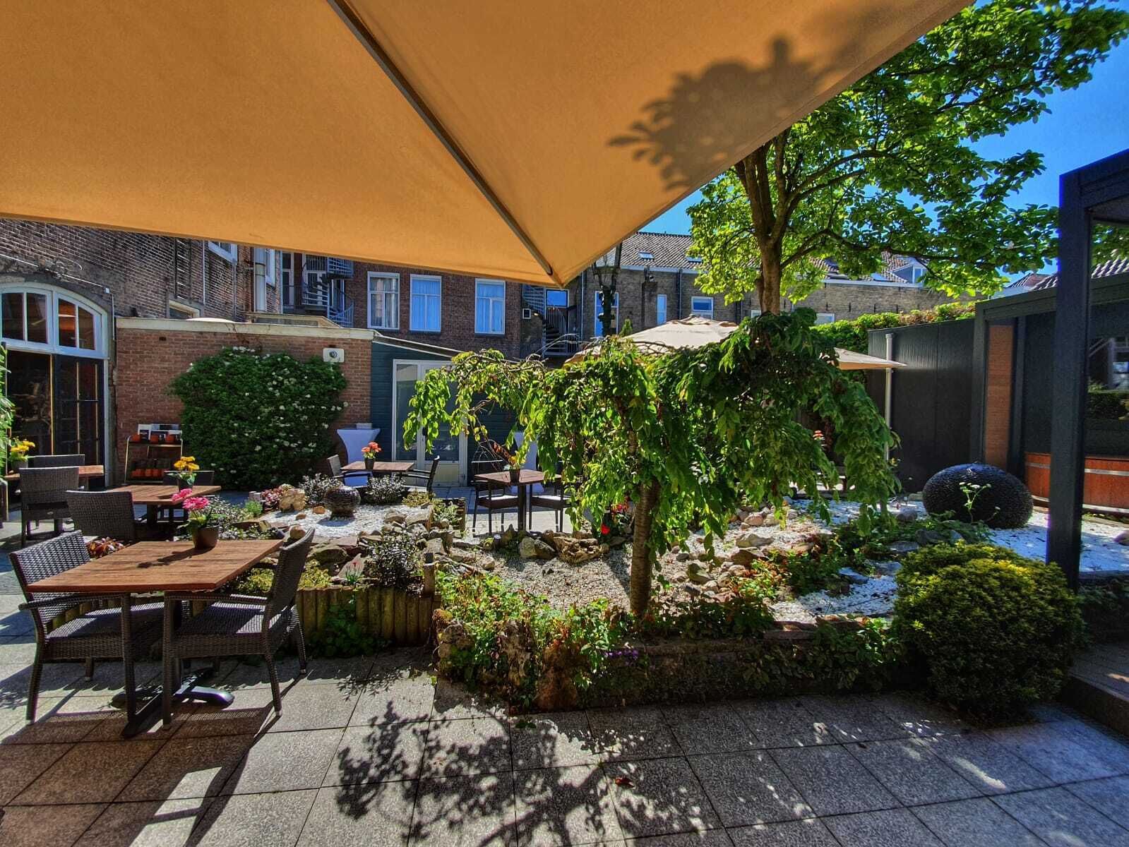 Une terrasse extérieure avec des tables et des chaises, entourée de verdure et avec un bâtiment en briques en arrière-plan.