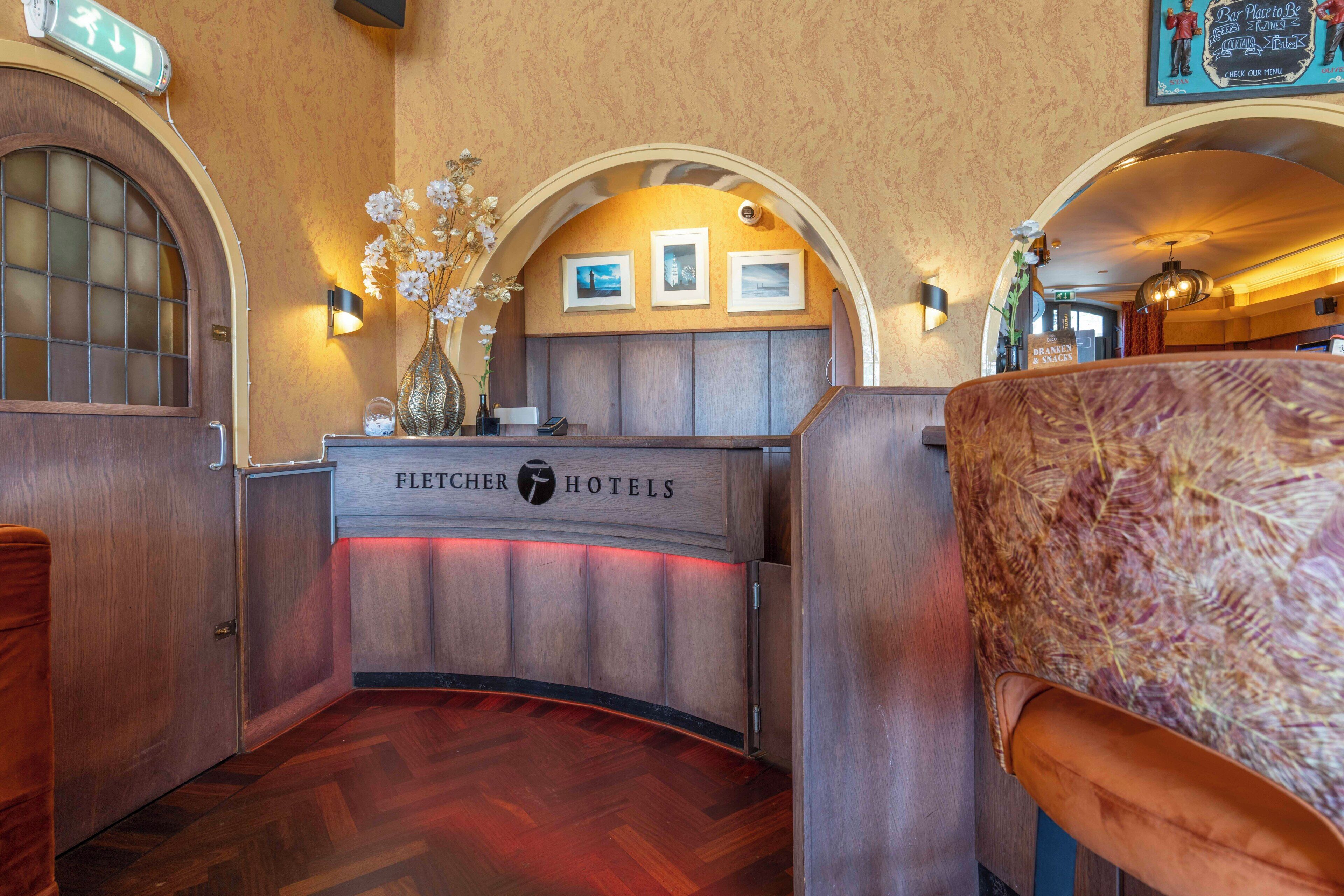 Ein Empfangsschalter mit Holzoptik, ein Schild mit der Aufschrift 'FLECHER HOTELS' und gerahmte Bilder an der Wand.